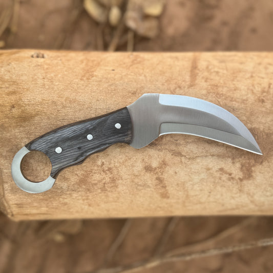 The Venomstrike Karambit Series featuring hand-forged high-carbon steel hawkbill blades, hardwood handles with integrated retention rings and brass pins, displayed with hand-tooled, hand-stitched leather pancake sheaths on a rugged rock and moss landscape, showcasing the knives’ curved karambit profiles, precision craftsmanship, and durable everyday-carry design available in Green, Orange, and Black finishes.