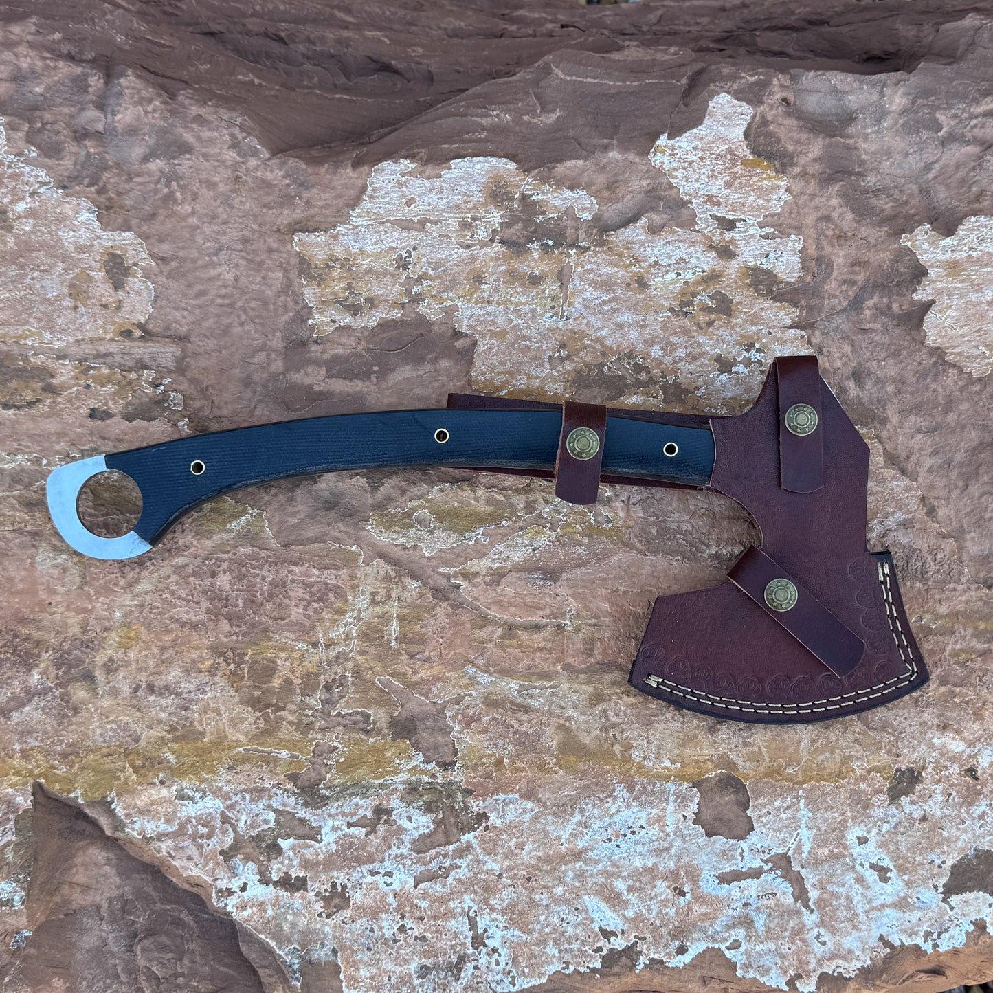 “A full-tang field axe with a stainless chopping head and a curved black micarta handle featuring brass hardware, photographed outdoors on stone. Designed for bushcraft, camping, chopping, and rugged outdoor field work.