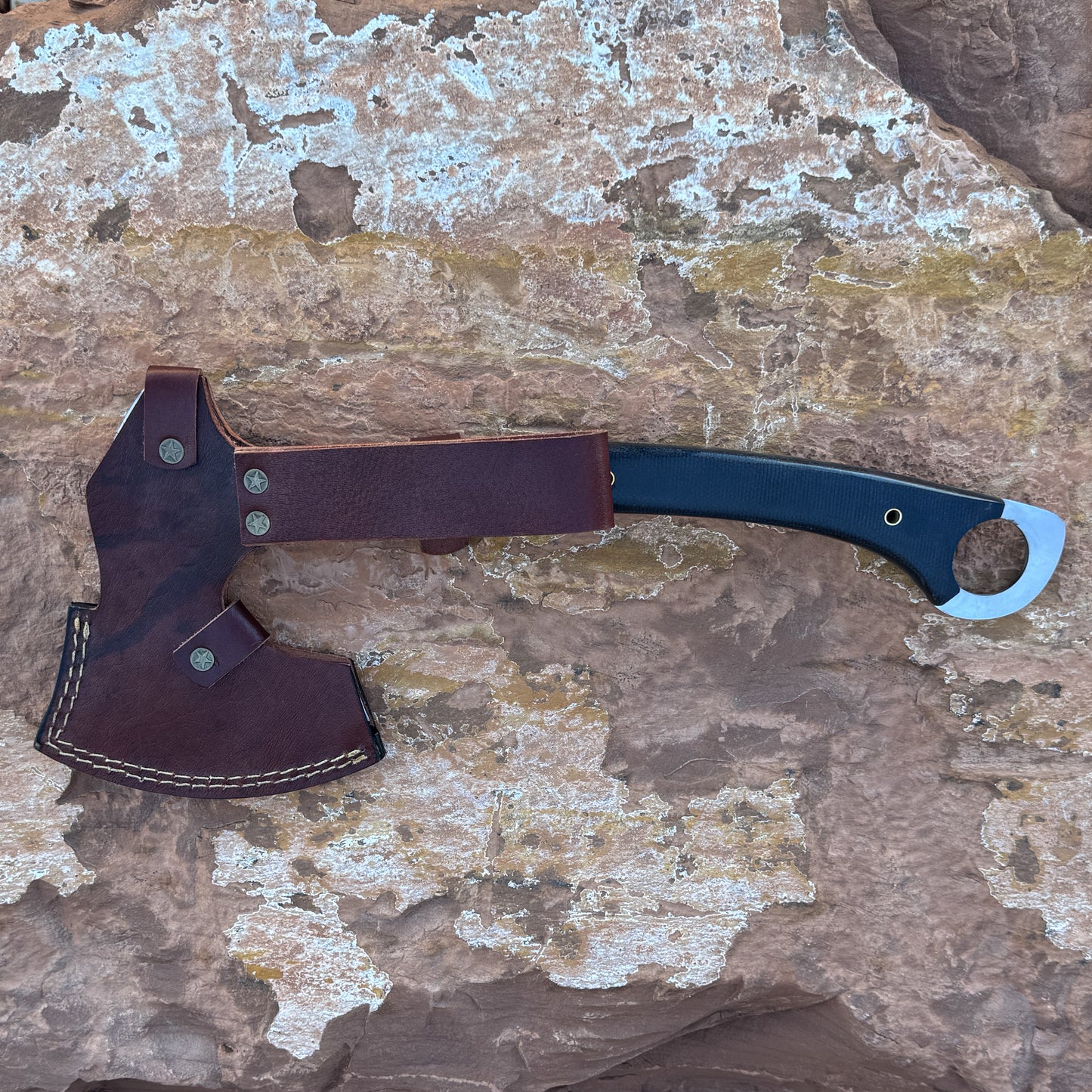 “A full-tang field axe with a stainless chopping head and a curved black micarta handle featuring brass hardware, photographed outdoors on stone. Designed for bushcraft, camping, chopping, and rugged outdoor field work.