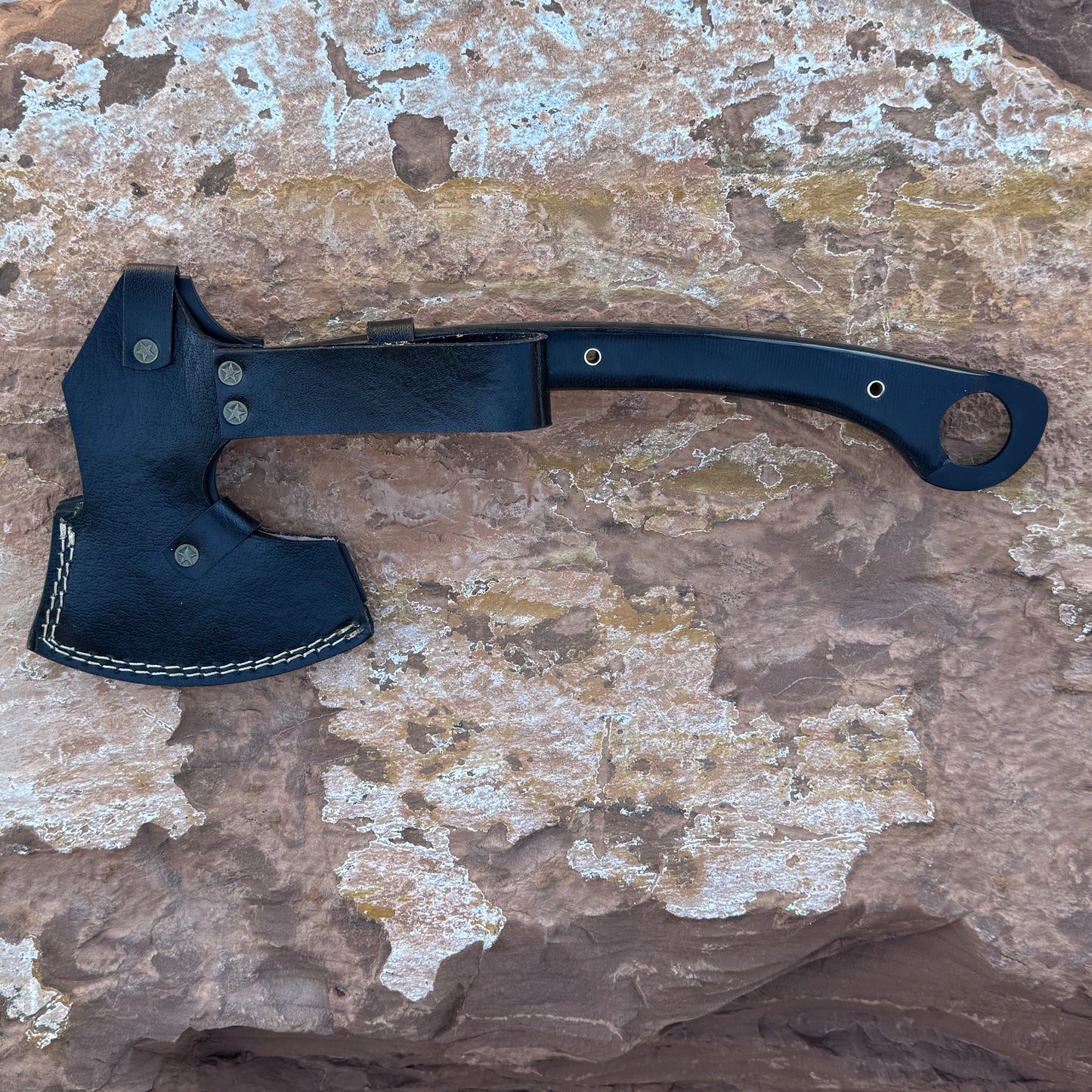 A handcrafted black leather sheath covering a 5160 steel tactical axe with a black micarta handle, displayed on desert rock. Built by Blue Spade Forge for outdoor tasks, chopping, and backcountry field use.
