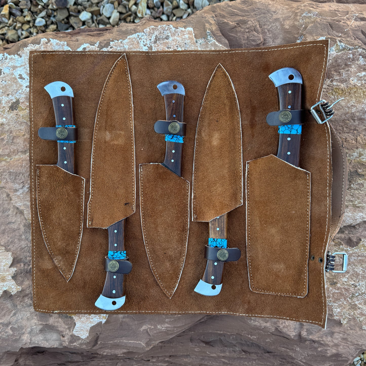 A premium suede-lined leather knife roll holding five Damascus kitchen knives with turquoise inlays and hardwood handles. The roll is displayed open on natural stone, showing stitched pockets and buckle closure.