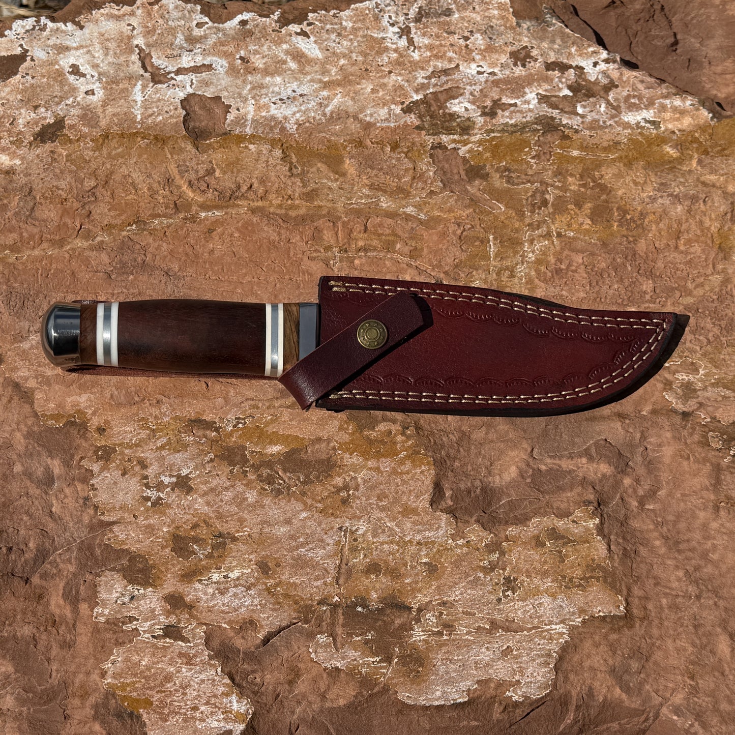 Frontier Talon Bowie knife with leather sheath showing full tang damascus blade and traditional wood handle on rugged stone background