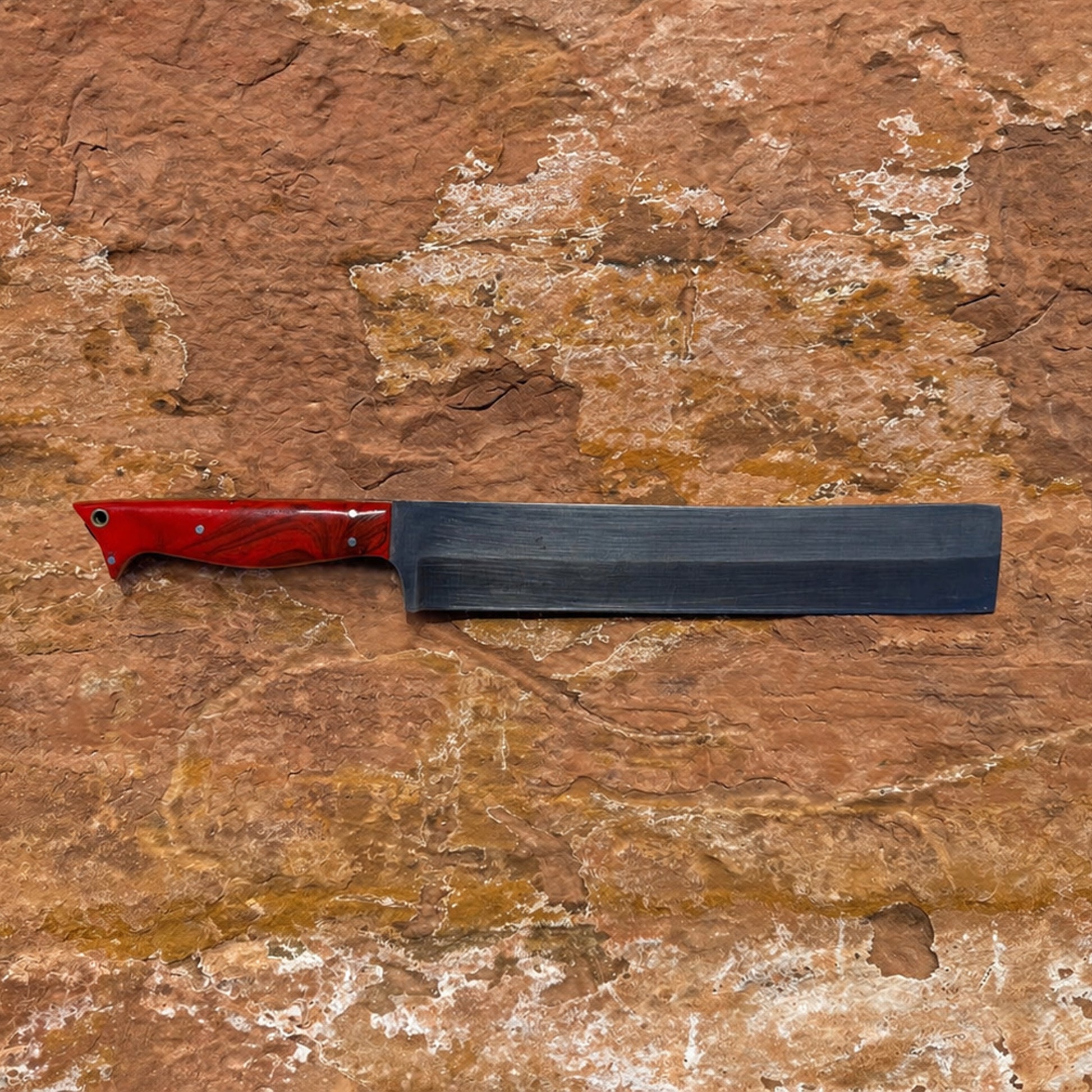 hand forged 1095 high carbon steel outdoor field chopper with red resin handle and leather sheath on natural rock surface
