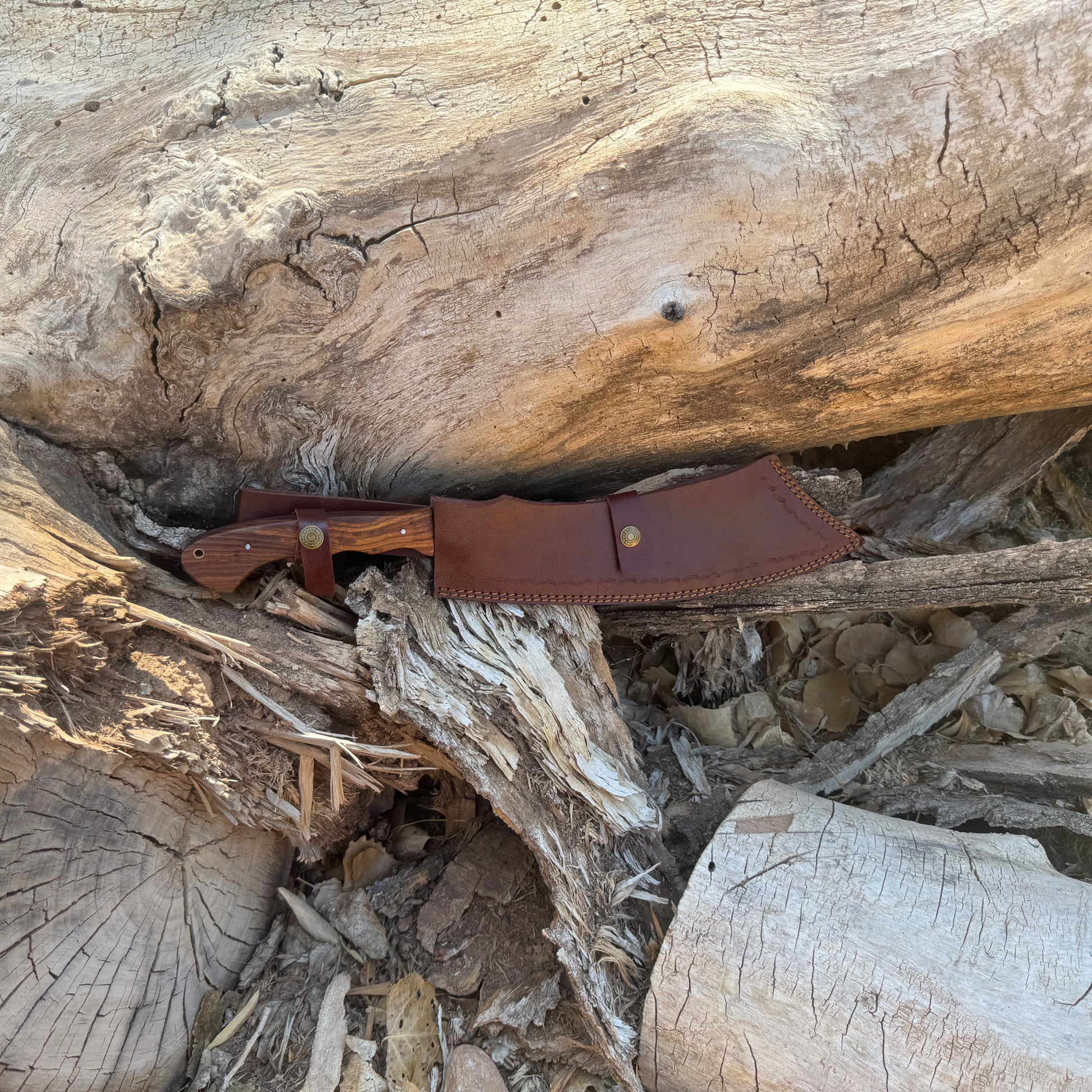 The Dreadfang machete featuring a hand-forged clip-point blade with a smooth walnut handle, displayed outdoors on a natural wooden surface to highlight its chopping power, precise piercing profile, rugged full-tang construction, and handcrafted Blue Spade Forge quality built for brush clearing and camp work.