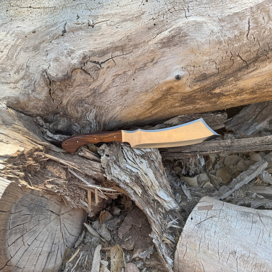 The Dreadfang machete featuring a hand-forged clip-point blade with a smooth walnut handle, displayed outdoors on a natural wooden surface to highlight its chopping power, precise piercing profile, rugged full-tang construction, and handcrafted Blue Spade Forge quality built for brush clearing and camp work.