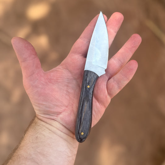 The Sentinel EDC knife featuring a hand-forged high-carbon-steel blade and an ergonomic ebony-wood handle with brass pins, shown beside its hand-tooled, double-stitched leather pancake sheath on a rugged rock and moss landscape, highlighting its durable everyday-carry design, refined hardwood grip, and dependable craftsmanship for daily or outdoor use.