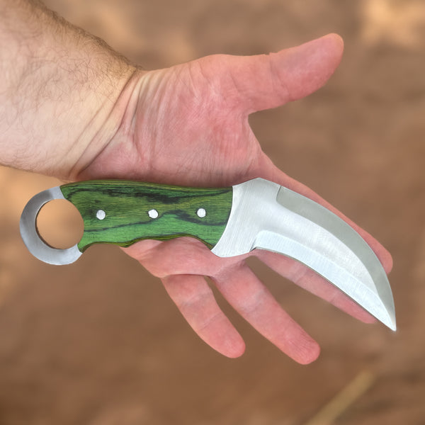 The Venomstrike Karambit Series featuring hand-forged high-carbon steel hawkbill blades, hardwood handles with integrated retention rings and brass pins, displayed with hand-tooled, hand-stitched leather pancake sheaths on a rugged rock and moss landscape, showcasing the knives’ curved karambit profiles, precision craftsmanship, and durable everyday-carry design available in Green, Orange, and Black finishes.