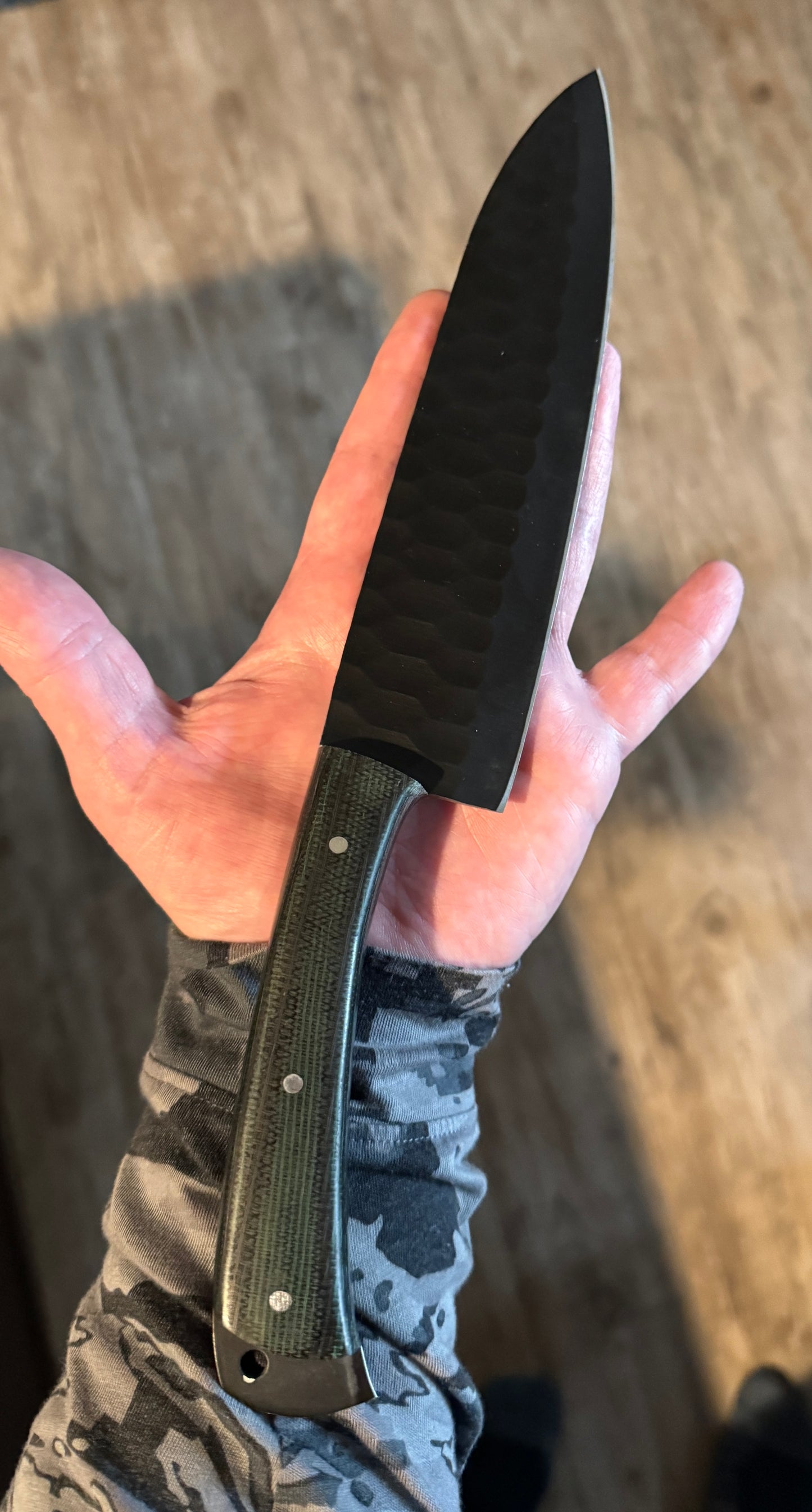 A powder-coated, hammered-finish full-tang chef’s knife with a contoured green-micarta handle, shown resting across the palm of a hand against a natural wood floor, highlighting the textured blade surface, ergonomic micarta grip, and durable handcrafted Blue Spade Forge design built for everyday culinary and outdoor cooking use.