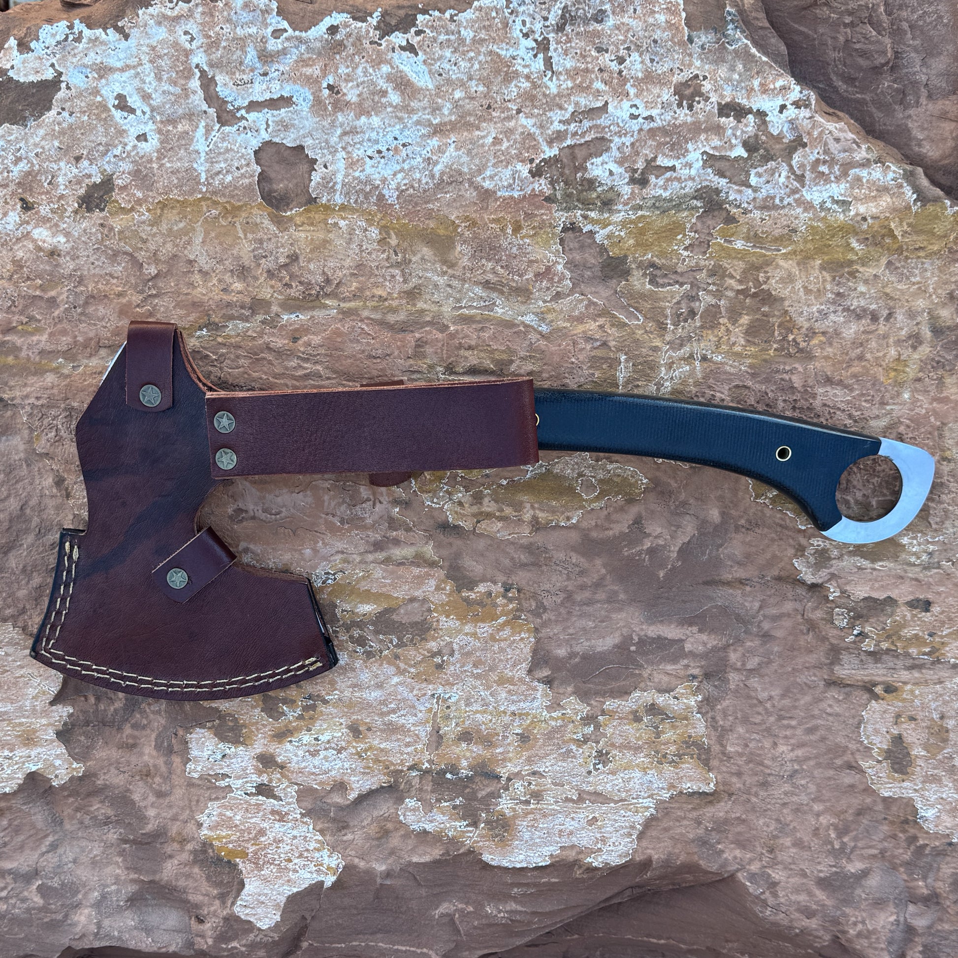“A full-tang field axe with a stainless chopping head and a curved black micarta handle featuring brass hardware, photographed outdoors on stone. Designed for bushcraft, camping, chopping, and rugged outdoor field work.