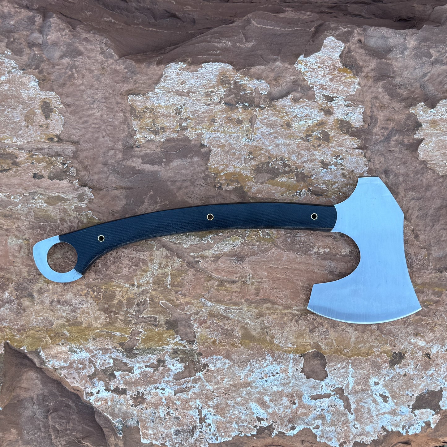 “A full-tang field axe with a stainless chopping head and a curved black micarta handle featuring brass hardware, photographed outdoors on stone. Designed for bushcraft, camping, chopping, and rugged outdoor field work.