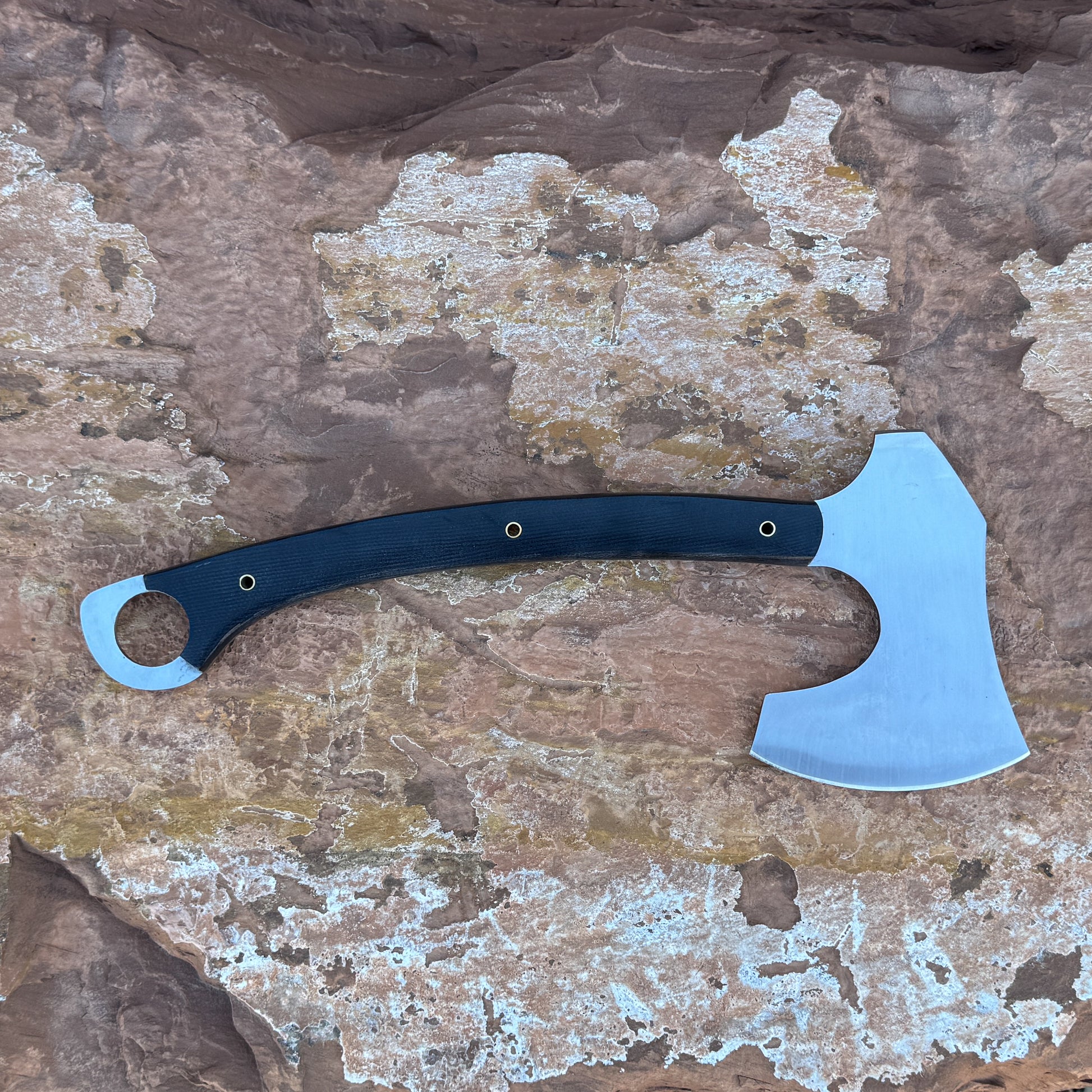 “A full-tang field axe with a stainless chopping head and a curved black micarta handle featuring brass hardware, photographed outdoors on stone. Designed for bushcraft, camping, chopping, and rugged outdoor field work.