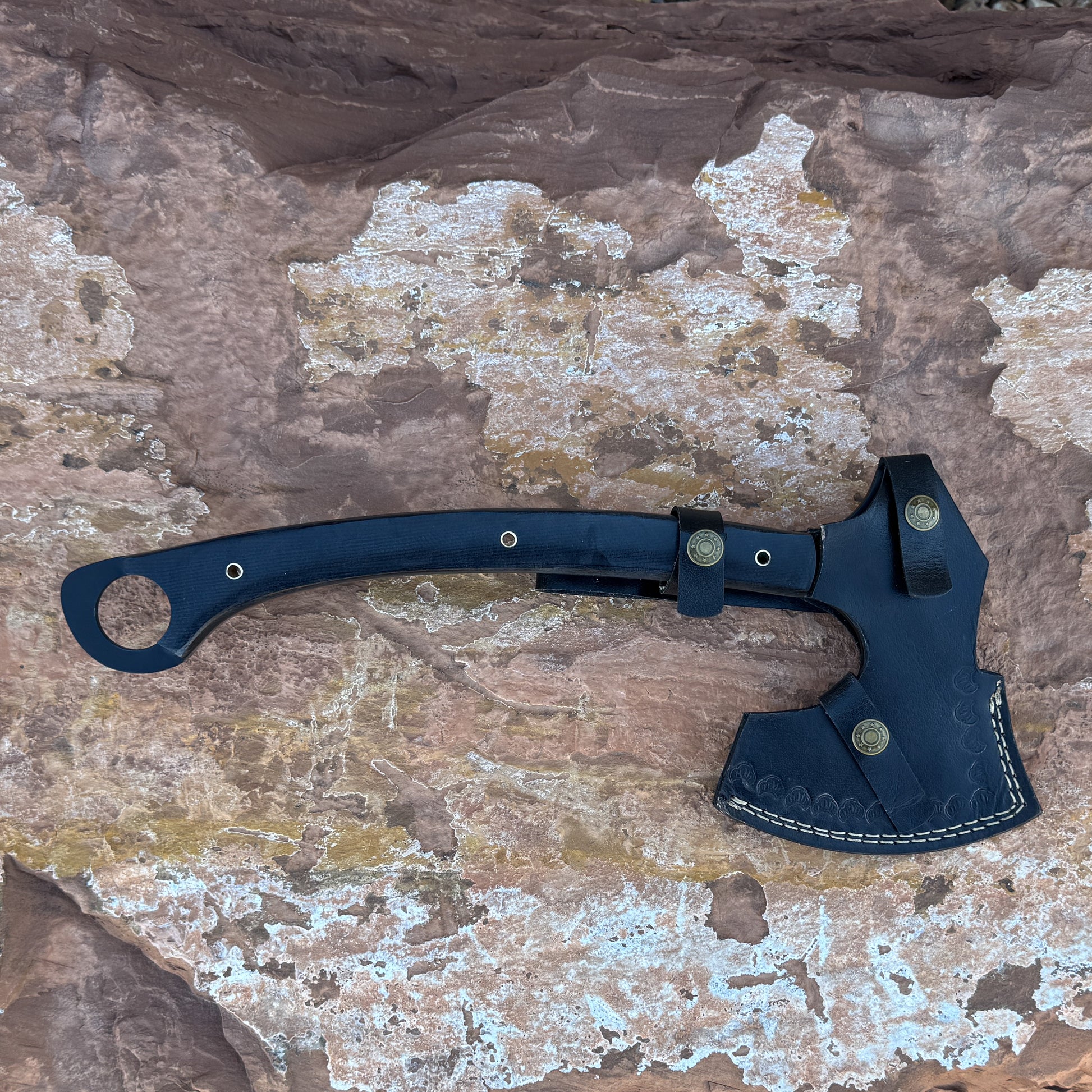 A handcrafted black leather sheath covering a 5160 steel tactical axe with a black micarta handle, displayed on desert rock. Built by Blue Spade Forge for outdoor tasks, chopping, and backcountry field use.