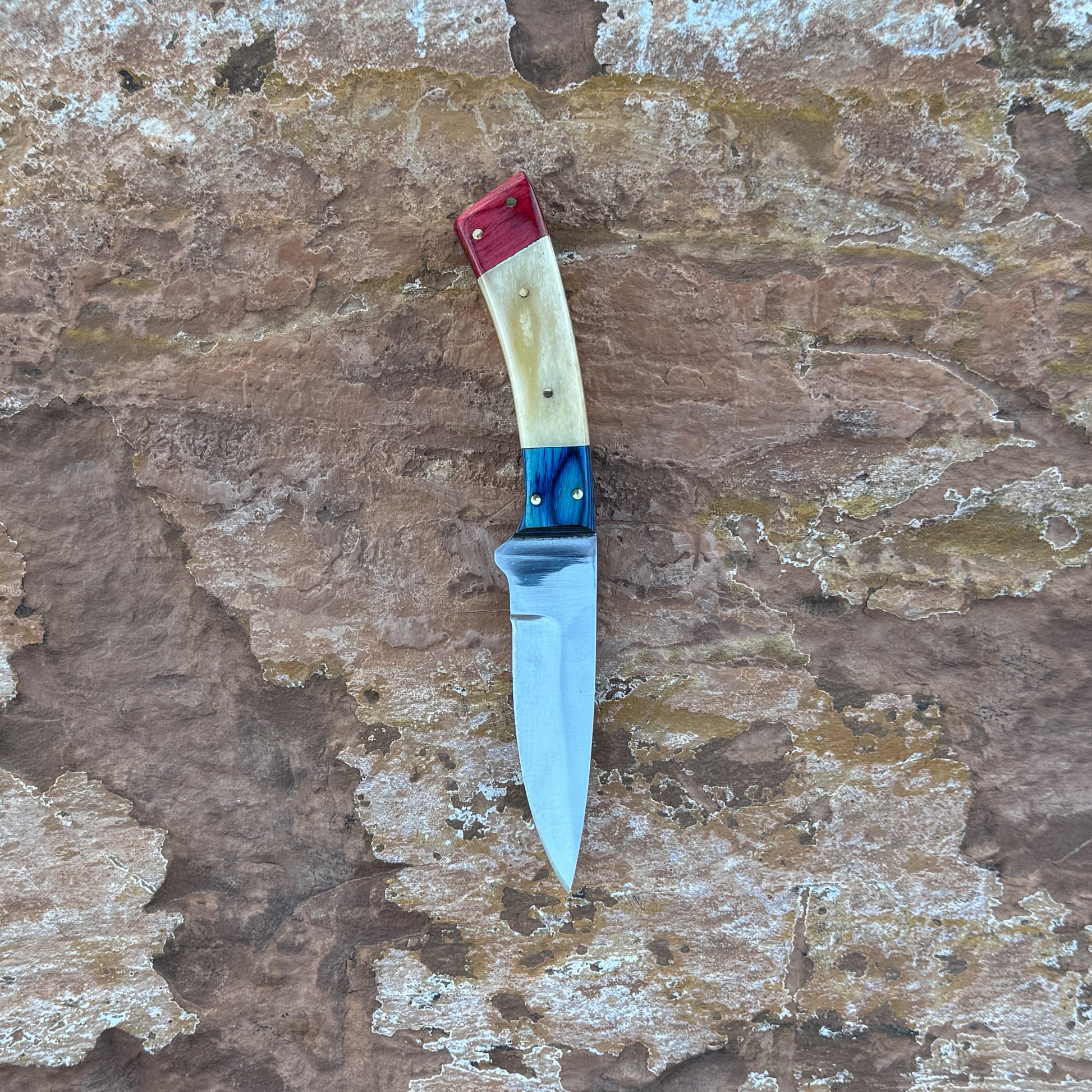 A fixed-blade knife with a red, natural camel bone, and blue dyed-wood tri-color handle stored in a hand-tooled black leather belt sheath, photographed on natural desert stone. Crafted by Blue Spade Forge for hunting, camping, and outdoor carry.