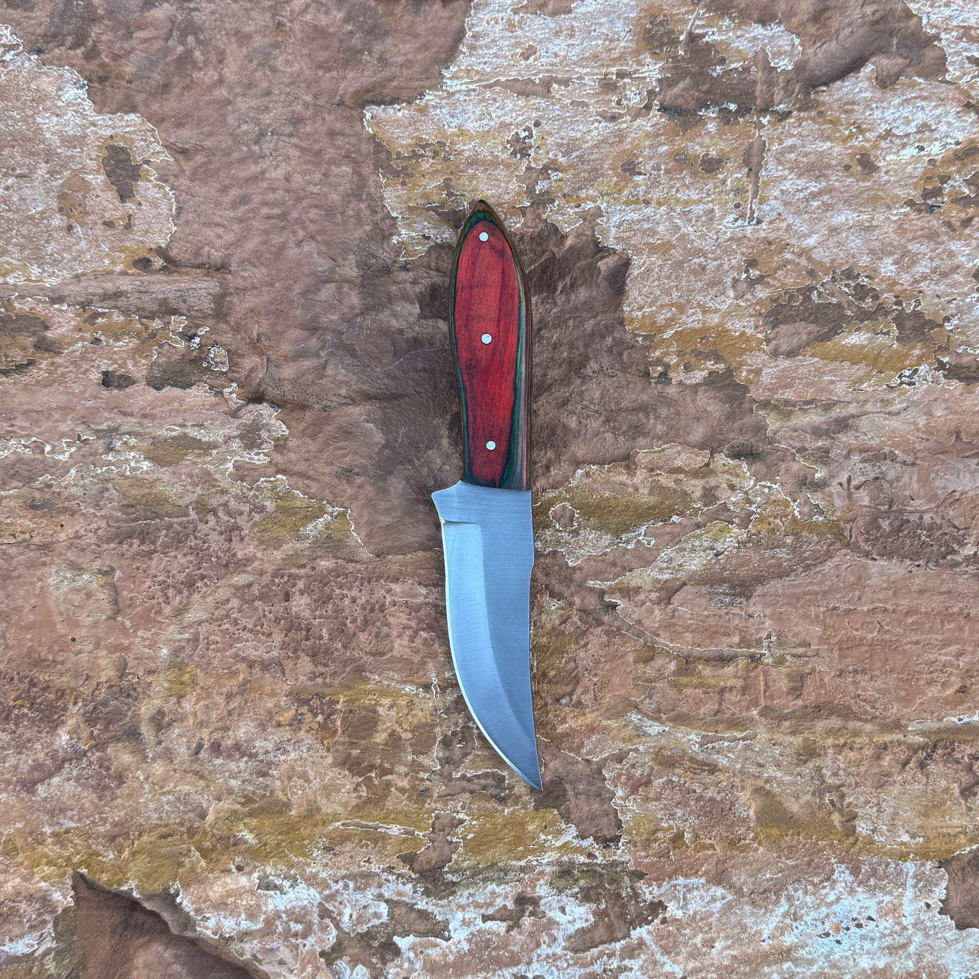 A full-tang fixed-blade knife made from 1095 high-carbon steel with a contoured red dyed-wood handle featuring rich grain patterns, shown with its hand-stitched brown leather sheath. Crafted by Blue Spade Forge for hunting, camping, carving, and rugged outdoor use.