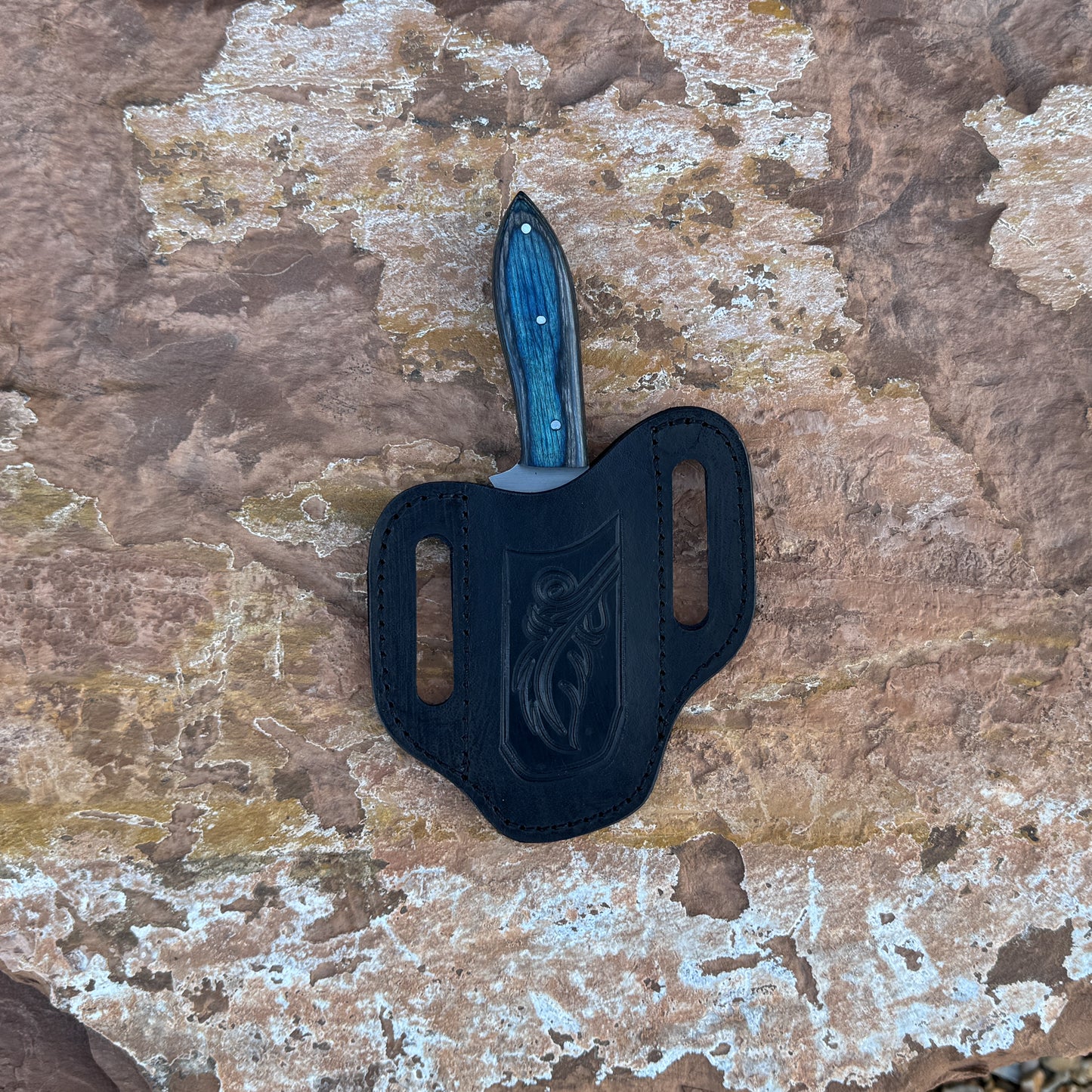 A full-tang clip-point hunting knife with a razor-sharp high-carbon steel blade and a blue-tinted wooden handle featuring bold grain patterns, shown with its hand-stitched leather sheath. Designed by Blue Spade Forge for hunting, field dressing, skinning, and rugged outdoor use.