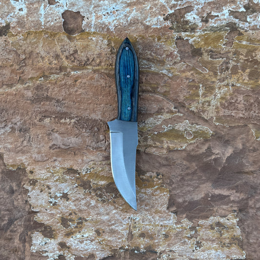 A full-tang clip-point hunting knife with a razor-sharp high-carbon steel blade and a blue-tinted wooden handle featuring bold grain patterns, shown with its hand-stitched leather sheath. Designed by Blue Spade Forge for hunting, field dressing, skinning, and rugged outdoor use.