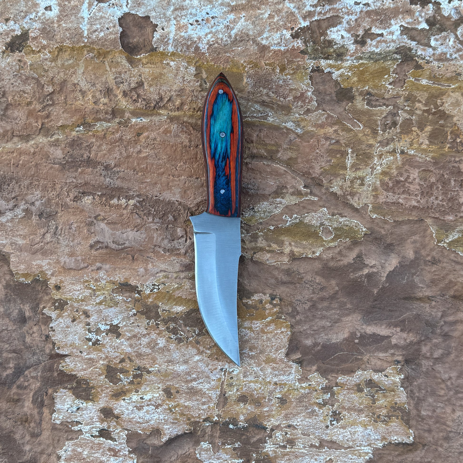 Blaze Creek Bowie knife with multicolored wood handle

Full-tang bowie knife on hand-stitched leather sheath

Clip-point blade close-up for slicing and skinning

Vivid blue, green, and red wood handle detail

Outdoor bowie knife displayed on natural wood

Triple-rivet handle construction on Blaze Creek

Blue Spade Forge Blaze Creek in natural lighting

Bowie knife and sheath arranged for product showcase