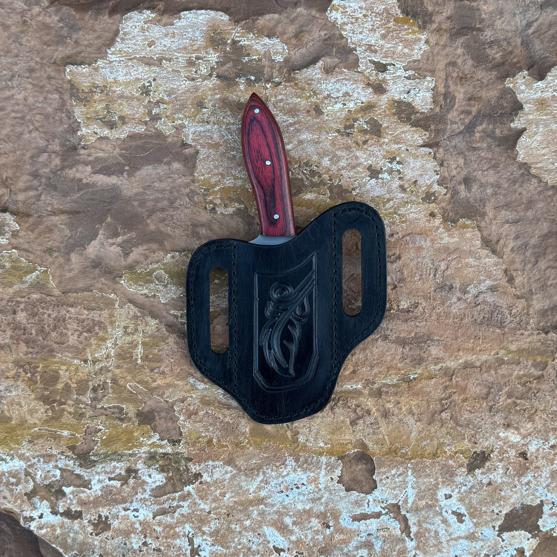 A full-tang fixed blade knife with a high-carbon steel blade and a deep red hardwood handle featuring bold grain patterns, shown with its hand-stitched brown leather sheath. Crafted by Blue Spade Forge for rugged outdoor tasks, carving, slicing, and field use.