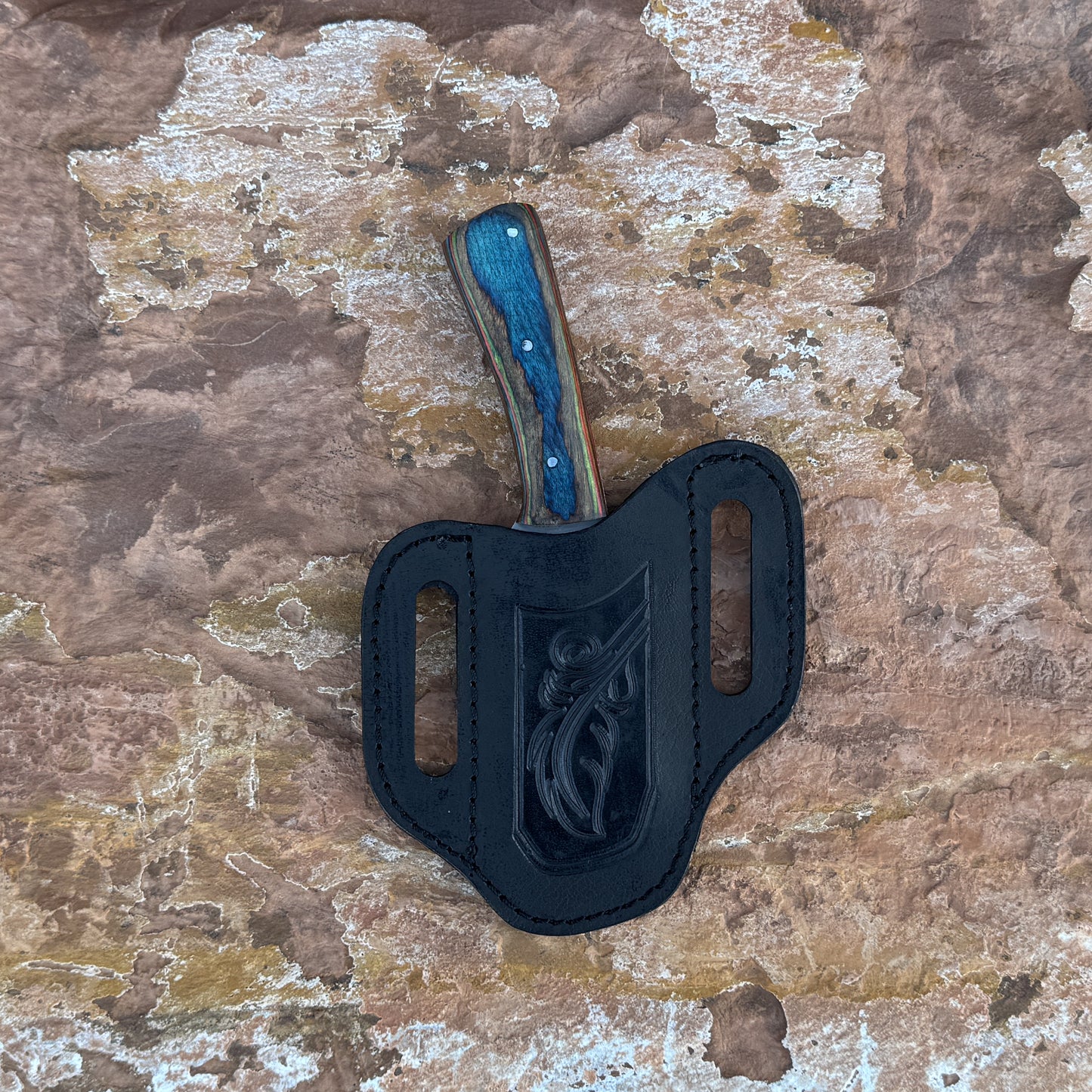 A full-tang 1095 high-carbon steel field knife with a contoured dyed-wood handle featuring bold color and rich grain patterns, shown with its hand-stitched brown leather sheath. Crafted by Blue Spade Forge for field use, camping, bushcraft, and rugged outdoor tasks