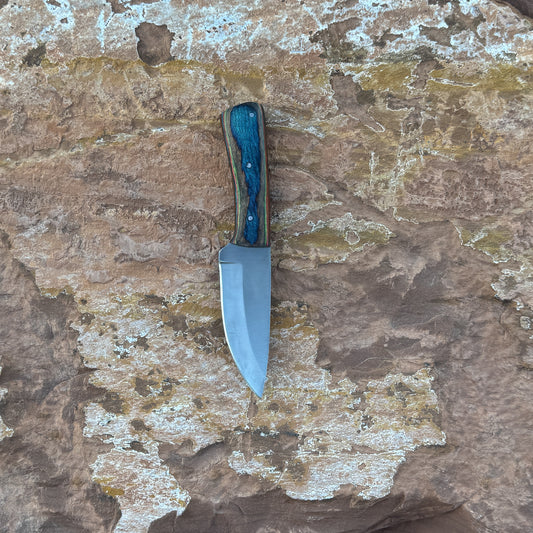 A full-tang 1095 high-carbon steel field knife with a contoured dyed-wood handle featuring bold color and rich grain patterns, shown with its hand-stitched brown leather sheath. Crafted by Blue Spade Forge for field use, camping, bushcraft, and rugged outdoor tasks