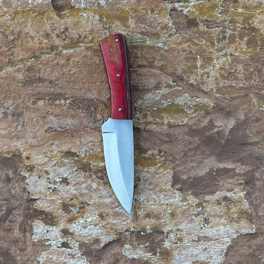 Crimson Forge Field Knife with red-grained hardwood handle

Drop-point satin blade displayed on leather sheath

Close-up of red hardwood handle with dual rivets

Field knife on natural wood background

Crimson Forge knife and hand-stitched sheath arranged together

Outdoor fixed blade in natural lighting

Blue Spade Forge Crimson Forge held in hand for scale

Drop-point blade and hardwood handle detail shot