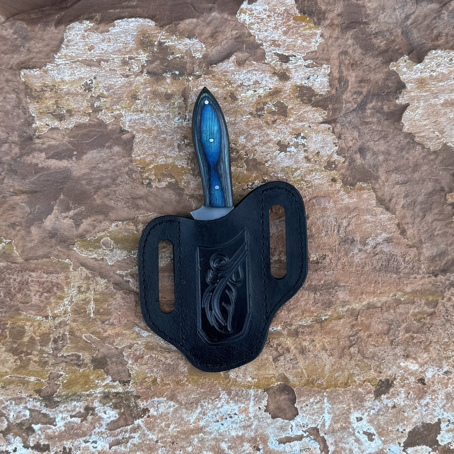 A full-tang upswept hunting knife with a satin-finished blade and deep-blue hardwood handle, reinforced with triple rivets. Shown alongside its hand-stitched leather sheath, crafted by Blue Spade Forge for field work, hunting, and rugged outdoor use.