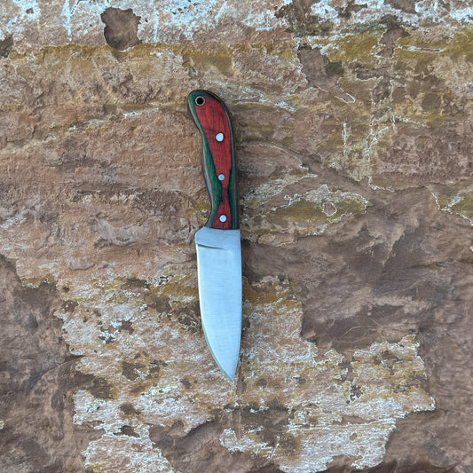 A full-tang stainless-steel field knife with a dyed-wood handle featuring rich grain patterns and vibrant coloration, shown with its handmade leather sheath. Designed by Blue Spade Forge for outdoor use, camping, and field tasks.