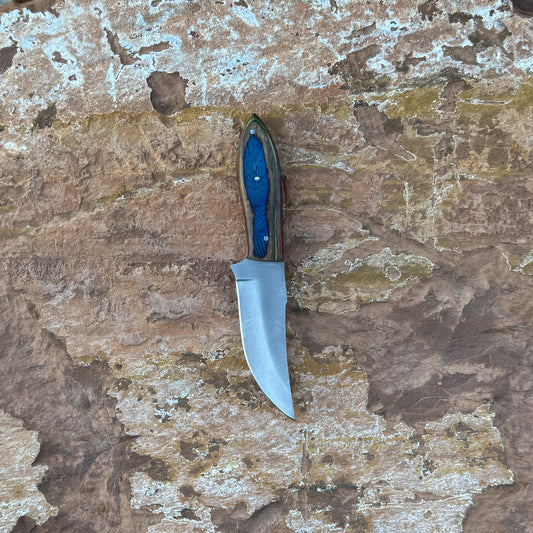 Blue Ridge Skinner knife with multi-tone hardwood handle

Sweeping clip-point blade displayed on leather sheath

Close-up of layered blue, red, green, and natural wood grain

Blue Ridge Skinner on natural wood background

Knife and hand-stitched leather sheath arranged together

Outdoor skinner knife in natural lighting

Blue Spade Forge Blue Ridge Skinner held in hand for scale

Detailed shot of clip-point blade and triple-riveted handle
