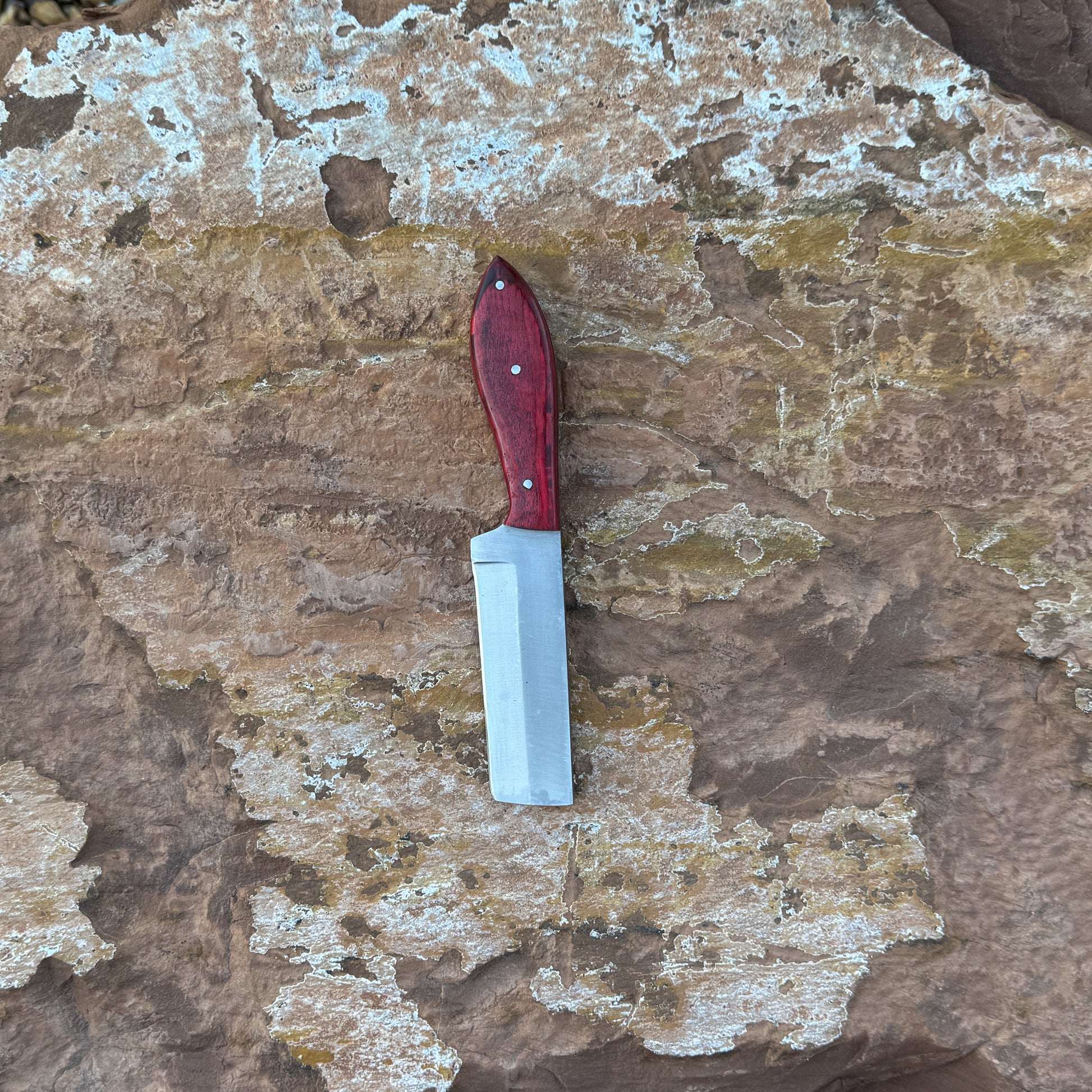 Scarlet Chop cowboy-style knife with squared 1095 blade

Close-up of red dyed hardwood handle

Compact cowboy knife shown with leather sheath

Blade detail showing squared chopping profile

Knife on rustic wood background

Scarlet Chop displayed with hand-stitched brown sheath

Blue Spade Forge Scarlet Chop held in hand for scale

Detail shot of blade edge and red handle grain