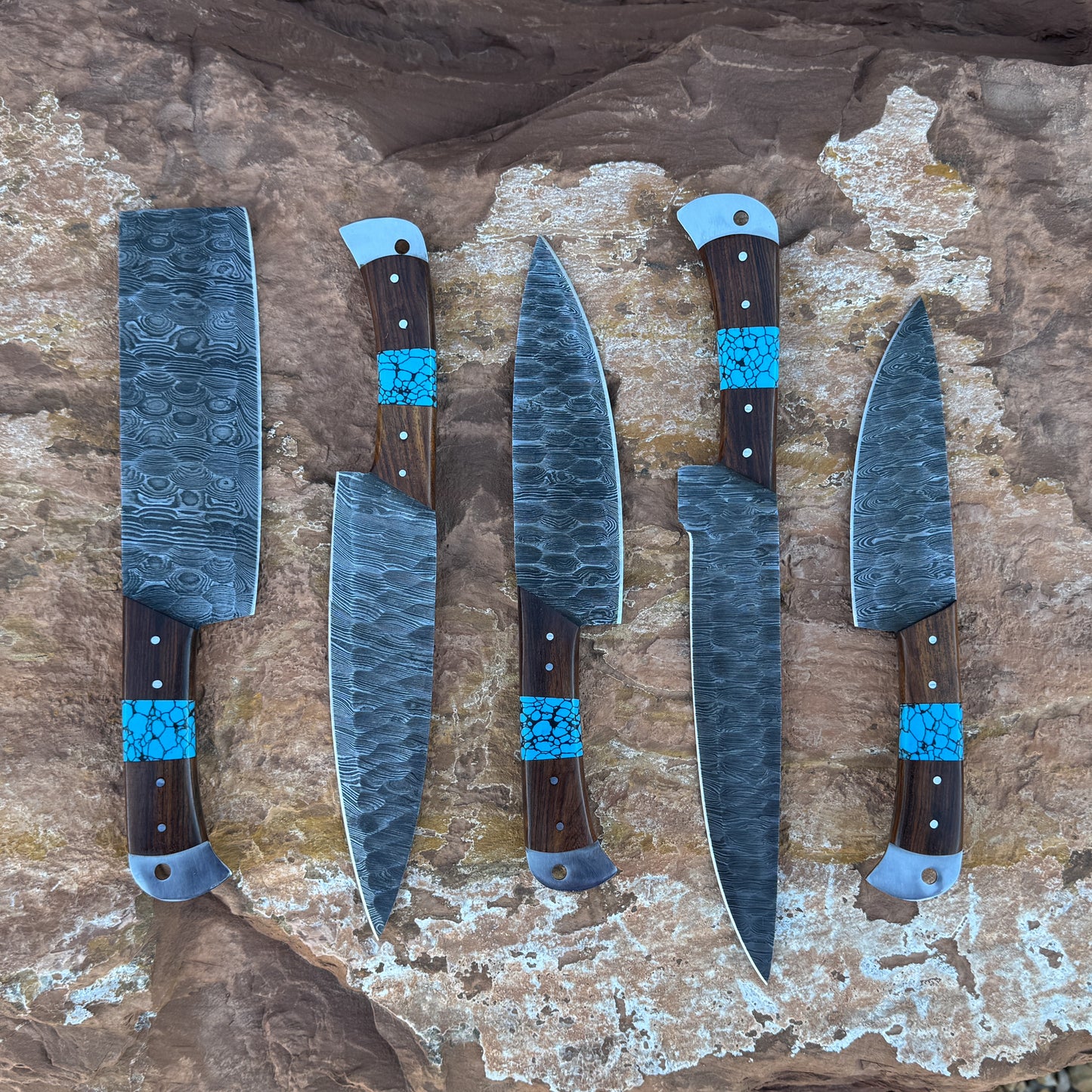 A five-piece handcrafted Damascus chef knife set with dark hardwood handles and vibrant turquoise stone inlays, displayed on natural desert rock. Includes a cleaver, butcher knife, chef knife, slicer, and utility knife
