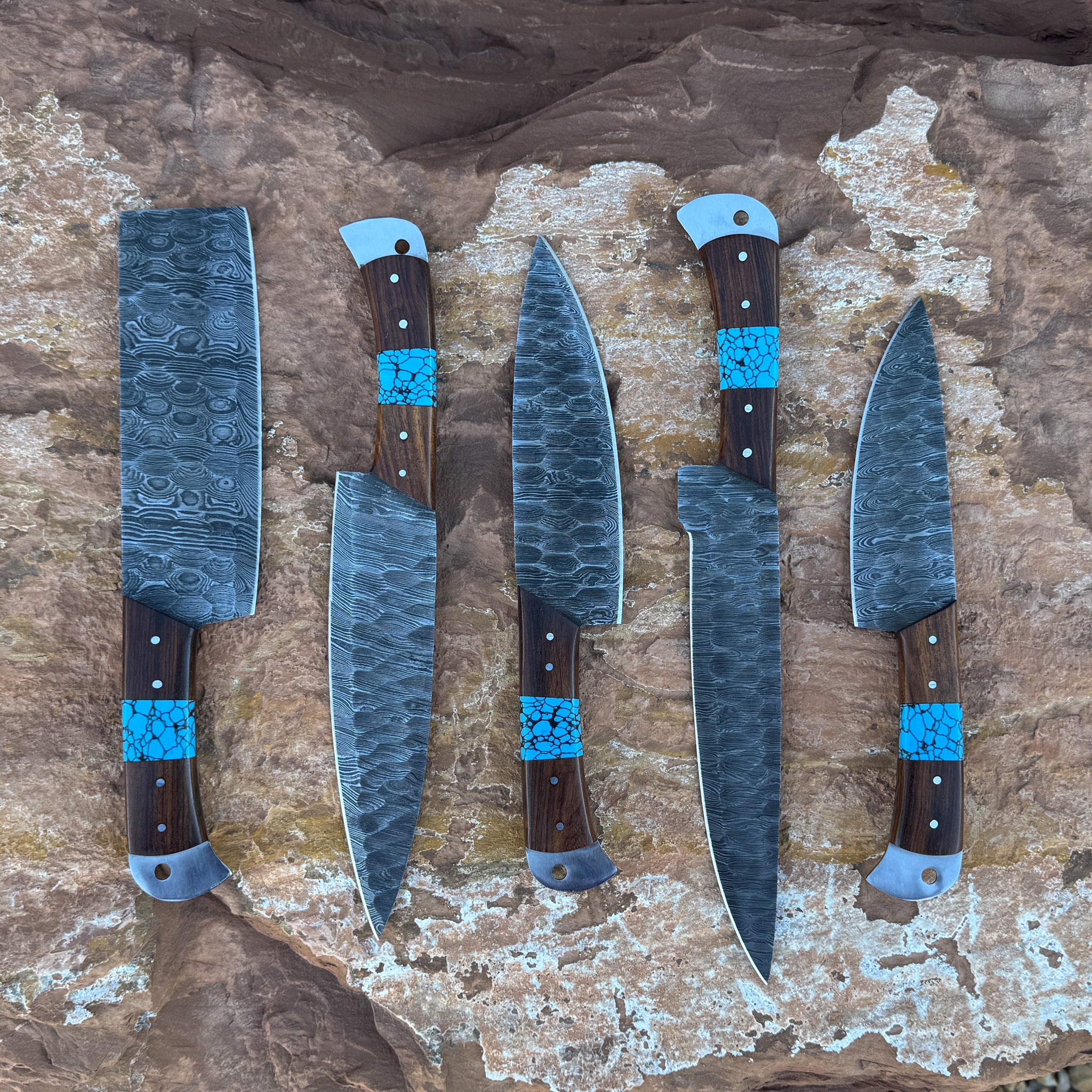 A five-piece handcrafted Damascus chef knife set with dark hardwood handles and vibrant turquoise stone inlays, displayed on natural desert rock. Includes a cleaver, butcher knife, chef knife, slicer, and utility knife