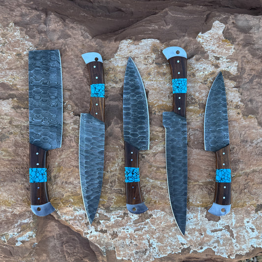 A five-piece handcrafted Damascus chef knife set with dark hardwood handles and vibrant turquoise stone inlays, displayed on natural desert rock. Includes a cleaver, butcher knife, chef knife, slicer, and utility knife
