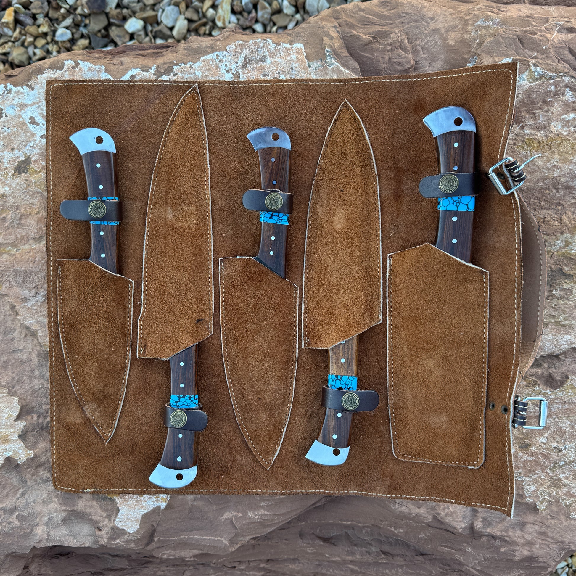 A premium suede-lined leather knife roll holding five Damascus kitchen knives with turquoise inlays and hardwood handles. The roll is displayed open on natural stone, showing stitched pockets and buckle closure.
