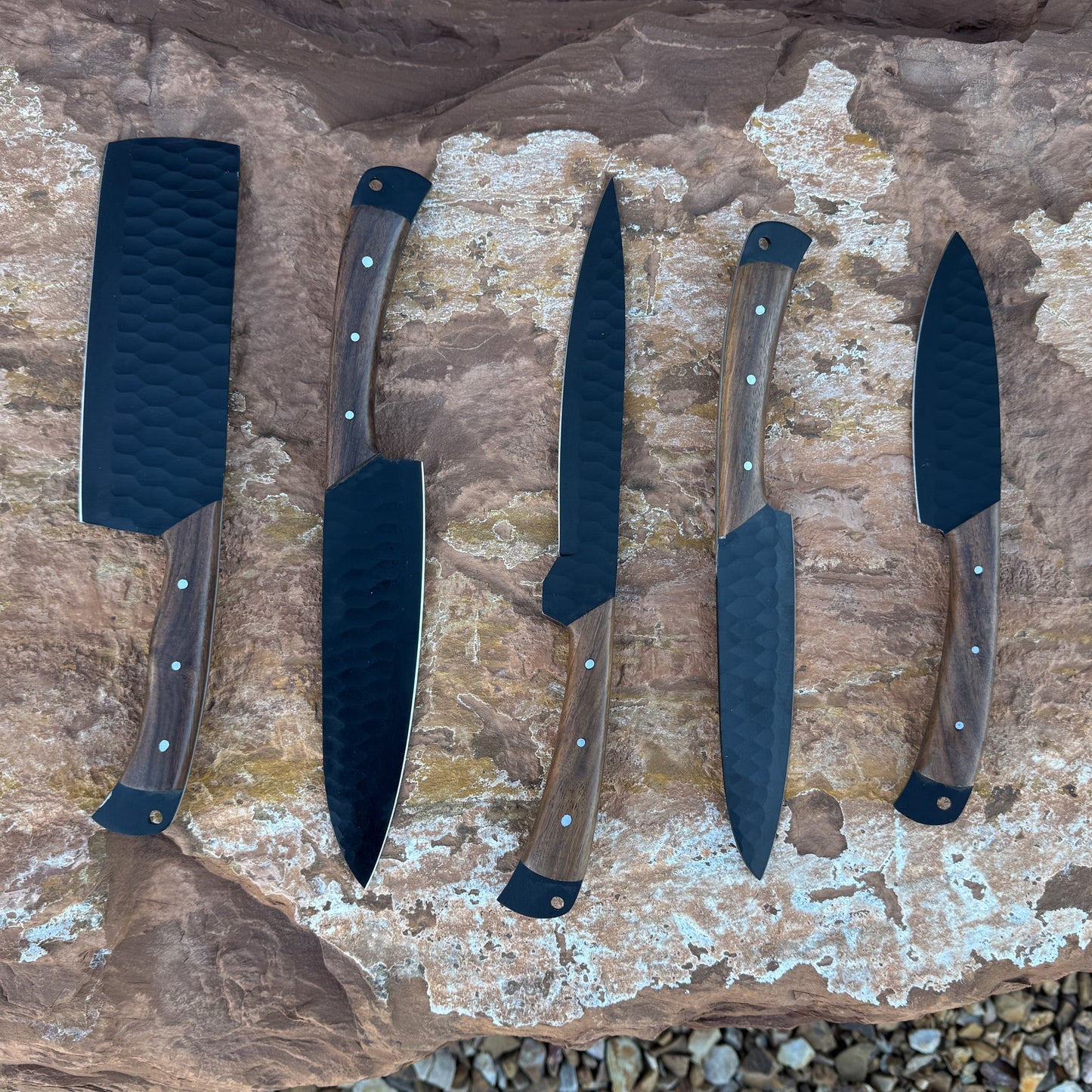 “A 5-piece chef knife set featuring matte black textured blades and dark hardwood handles displayed on natural stone. Includes a paring knife, utility knife, slicer, chef knife, and cleaver.
