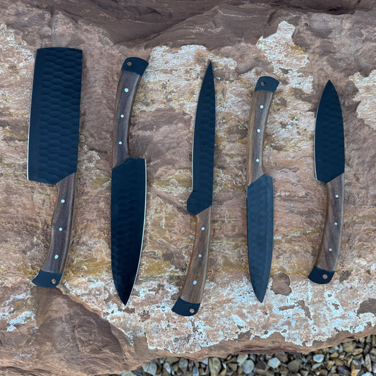 “A 5-piece chef knife set featuring matte black textured blades and dark hardwood handles displayed on natural stone. Includes a paring knife, utility knife, slicer, chef knife, and cleaver.