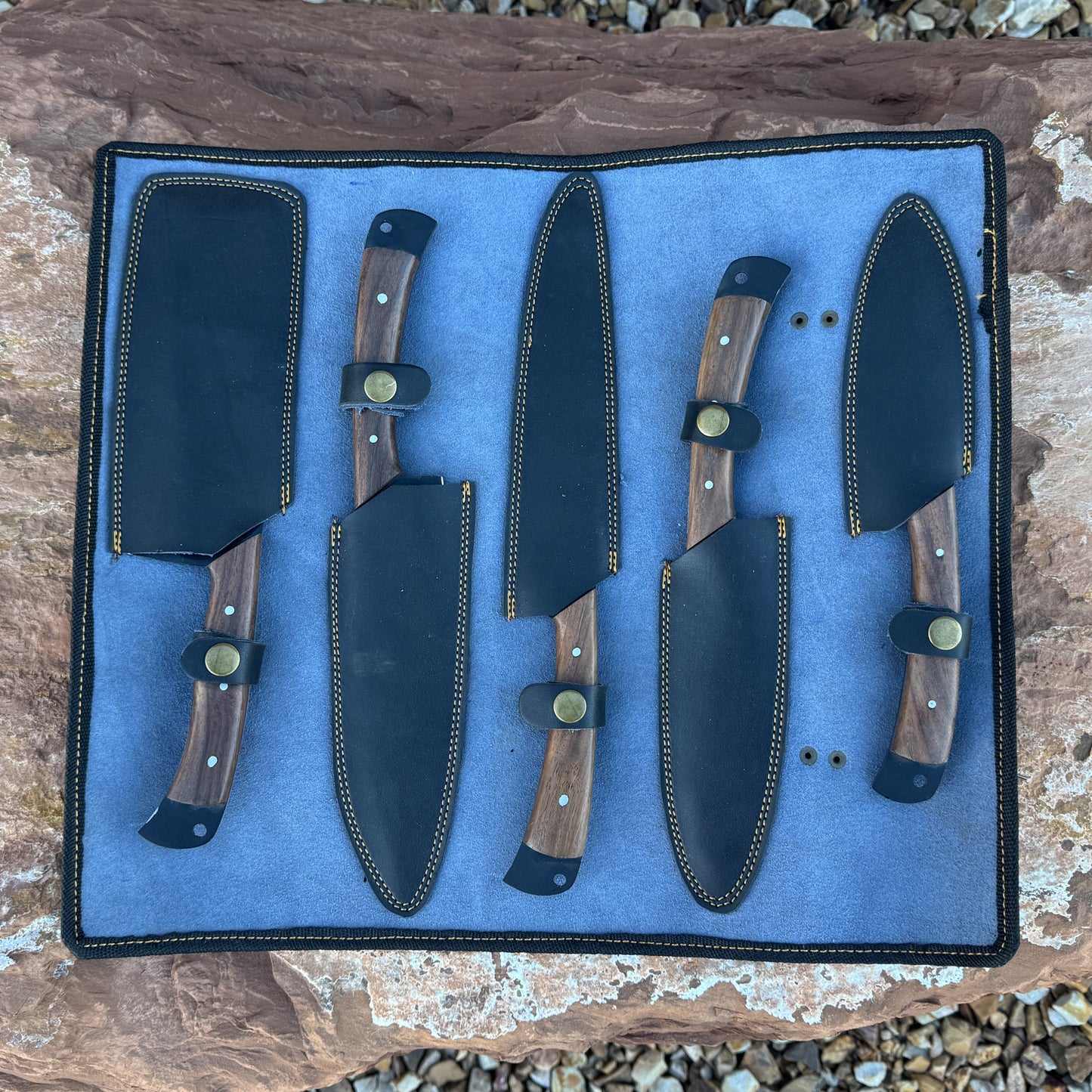 “A 5-piece chef knife set featuring matte black textured blades and dark hardwood handles displayed on natural stone. Includes a paring knife, utility knife, slicer, chef knife, and cleaver.