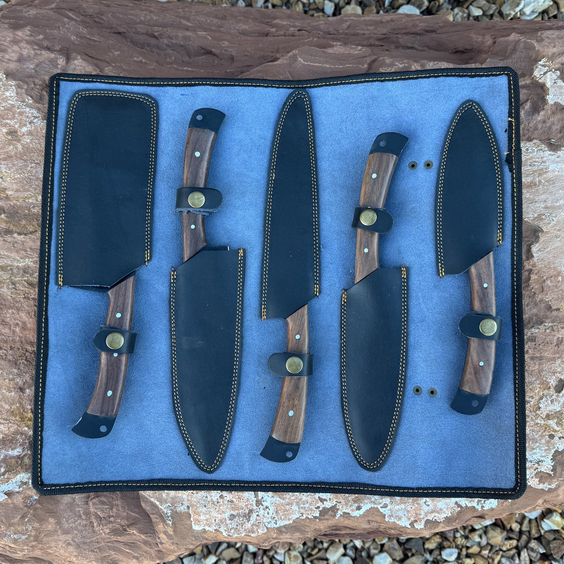 “A 5-piece chef knife set featuring matte black textured blades and dark hardwood handles displayed on natural stone. Includes a paring knife, utility knife, slicer, chef knife, and cleaver.