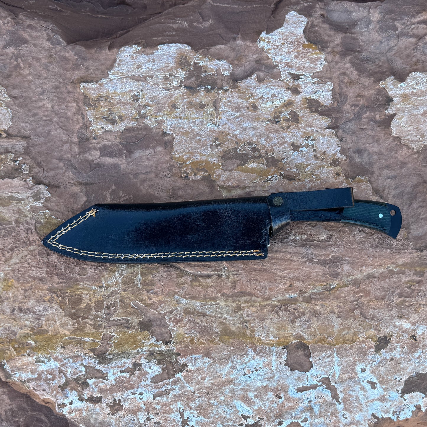 5160 high-carbon steel chef knife with a black powder-coated blade resting in a hand-stitched black leather sheath, featuring a contoured micarta handle and full-tang construction, photographed on a natural stone background