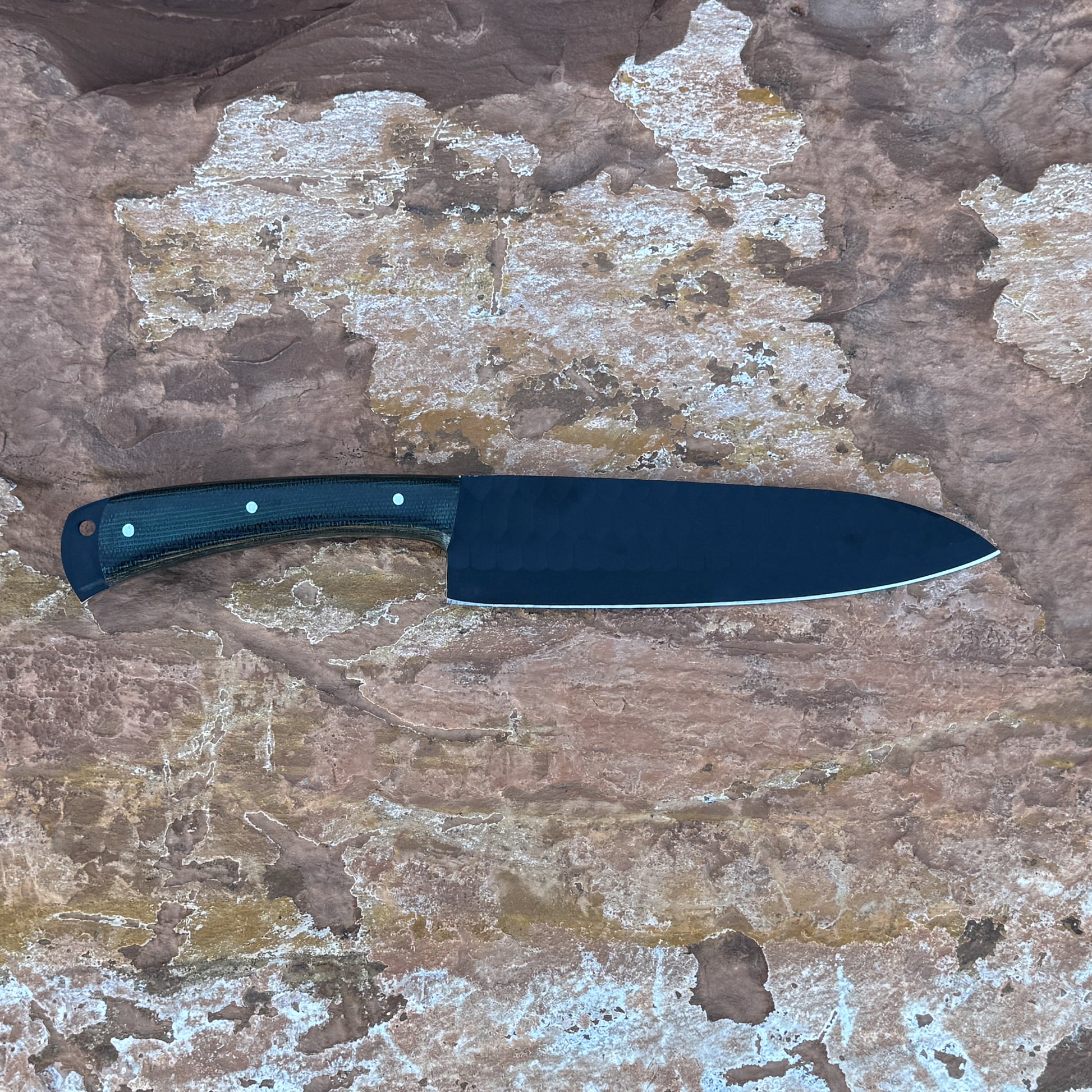 5160 high-carbon chef knife with black powder-coated blade, razor-sharp cutting edge, contoured micarta handle, and balanced profile designed for slicing, chopping, and professional kitchen use, shown unsheathed on textured stone surface