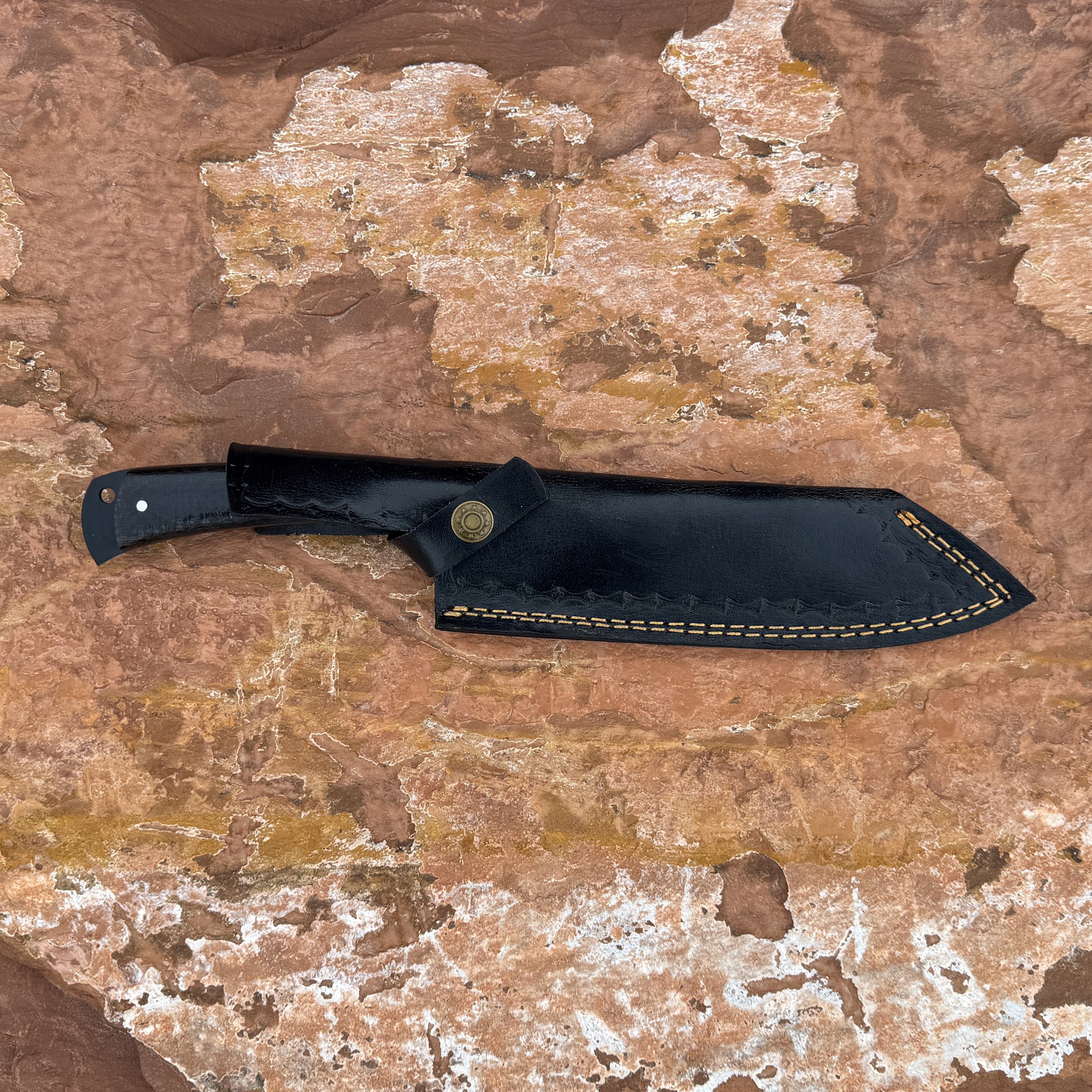 5160 high-carbon steel chef knife with a black powder-coated blade resting in a hand-stitched black leather sheath, featuring a contoured micarta handle and full-tang construction, photographed on a natural stone background