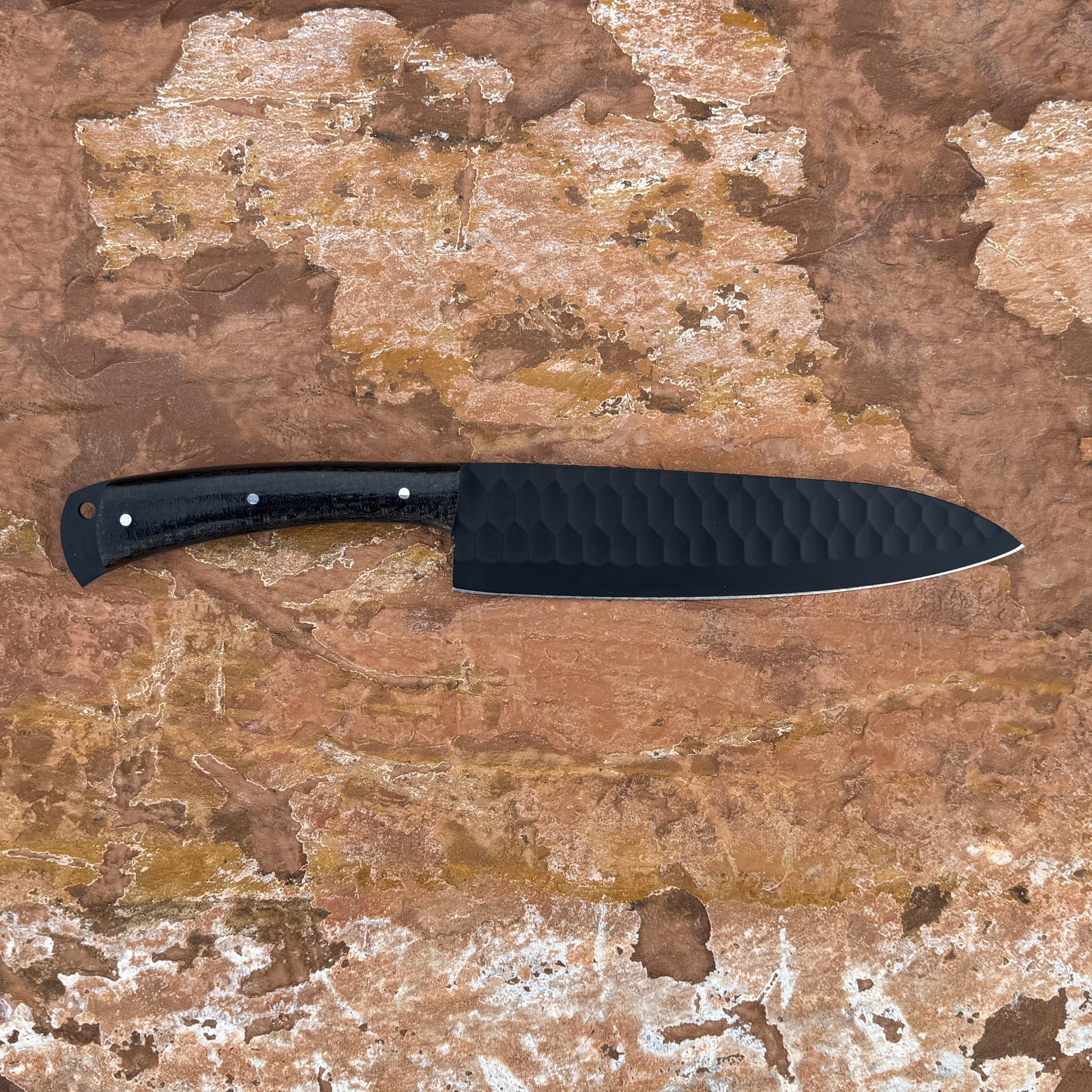 5160 high-carbon chef knife with black powder-coated blade, razor-sharp cutting edge, contoured micarta handle, and balanced profile designed for slicing, chopping, and professional kitchen use, shown unsheathed on textured stone surface