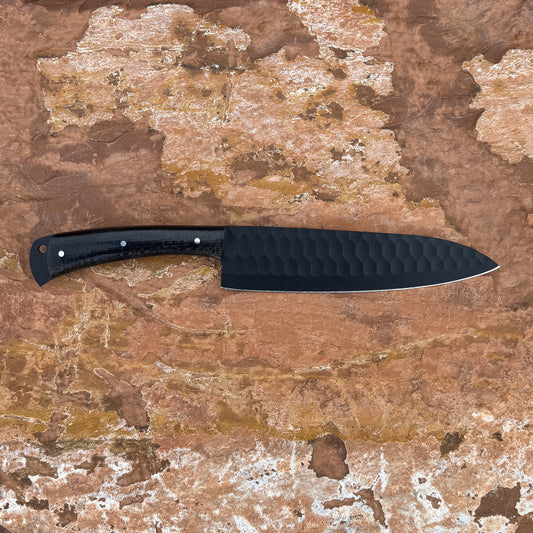 5160 high-carbon chef knife with black powder-coated blade, razor-sharp cutting edge, contoured micarta handle, and balanced profile designed for slicing, chopping, and professional kitchen use, shown unsheathed on textured stone surface