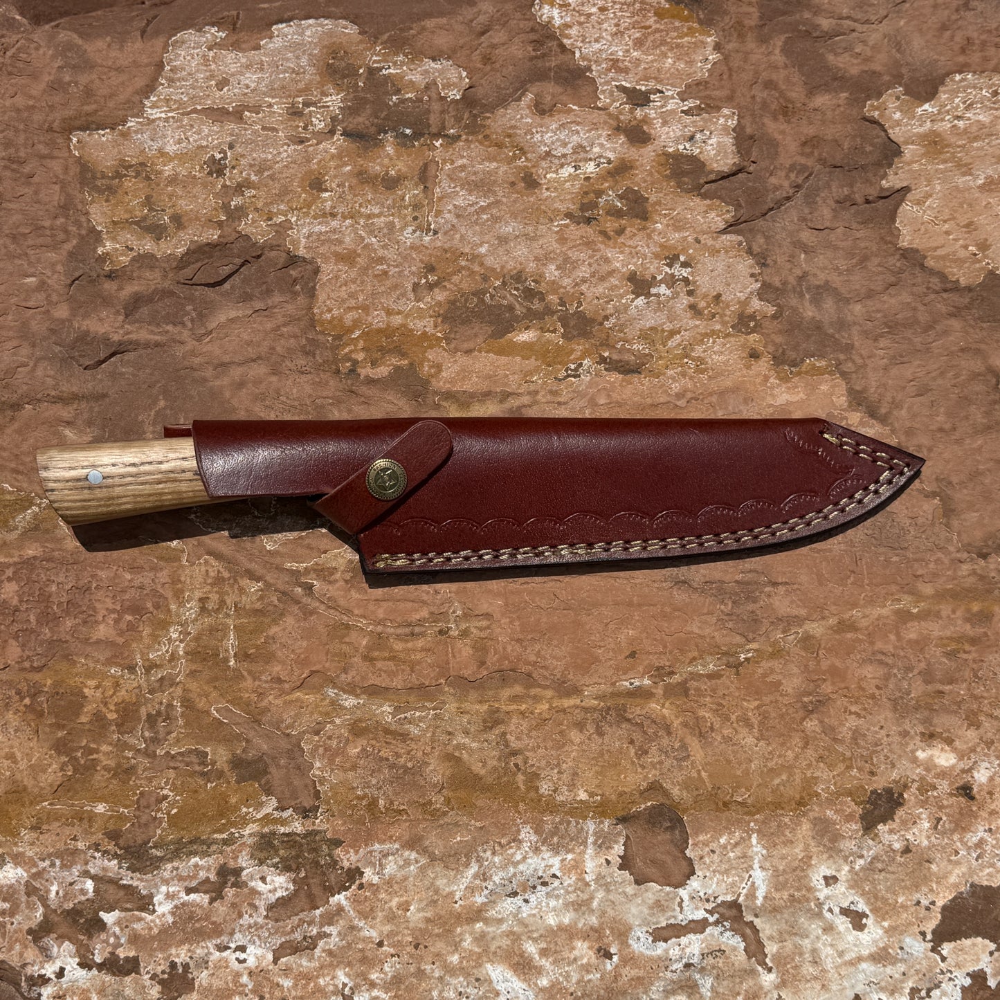 Hand-forged Blue Spade Forge Timberline Damascus chef knife featuring layered Damascus steel blade, hardwood handle with brass pins, and handcrafted brown leather sheath displayed on natural stone background.