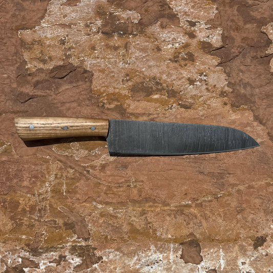 Hand-forged Blue Spade Forge Timberline Damascus chef knife featuring layered Damascus steel blade, hardwood handle with brass pins, and handcrafted brown leather sheath displayed on natural stone background.