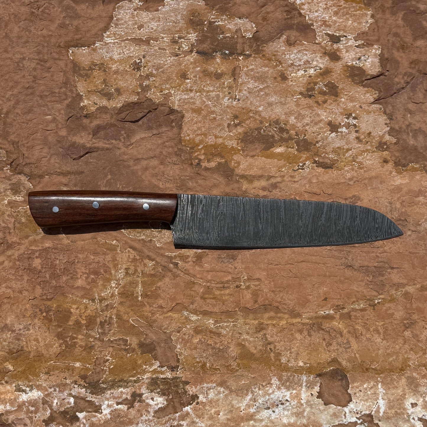 Hand-forged Blue Spade Forge Timberline Damascus chef knife featuring layered Damascus steel blade, hardwood handle with brass pins, and handcrafted brown leather sheath displayed on natural stone background.