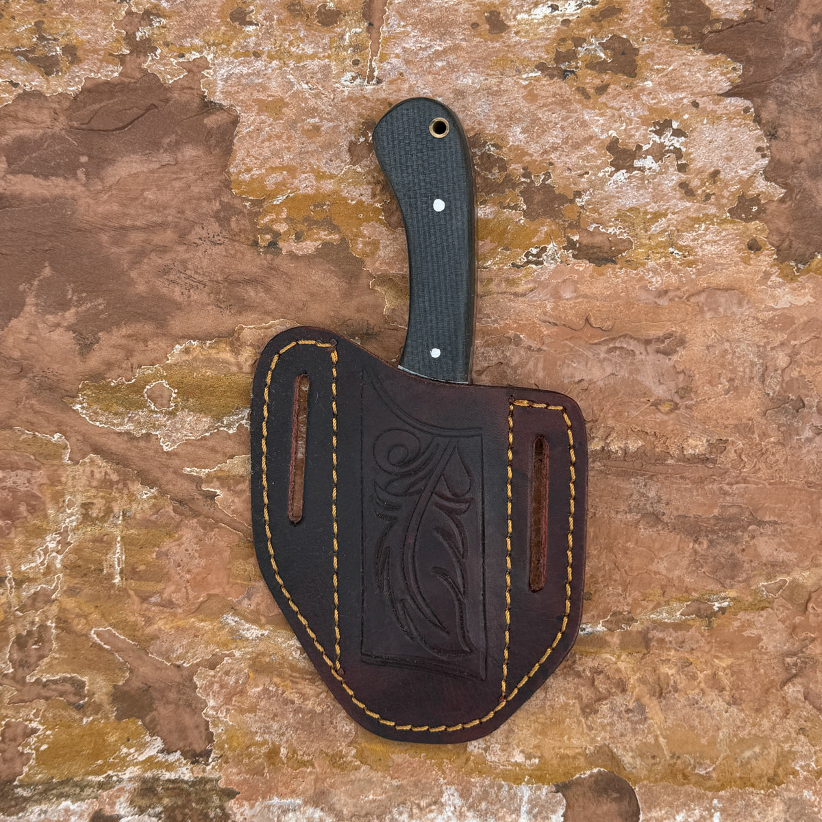 A dark brown leather knife sheath with tan stitching and an embossed leaf design, holding a black-handled knife, displayed against a textured brown and tan stone background.