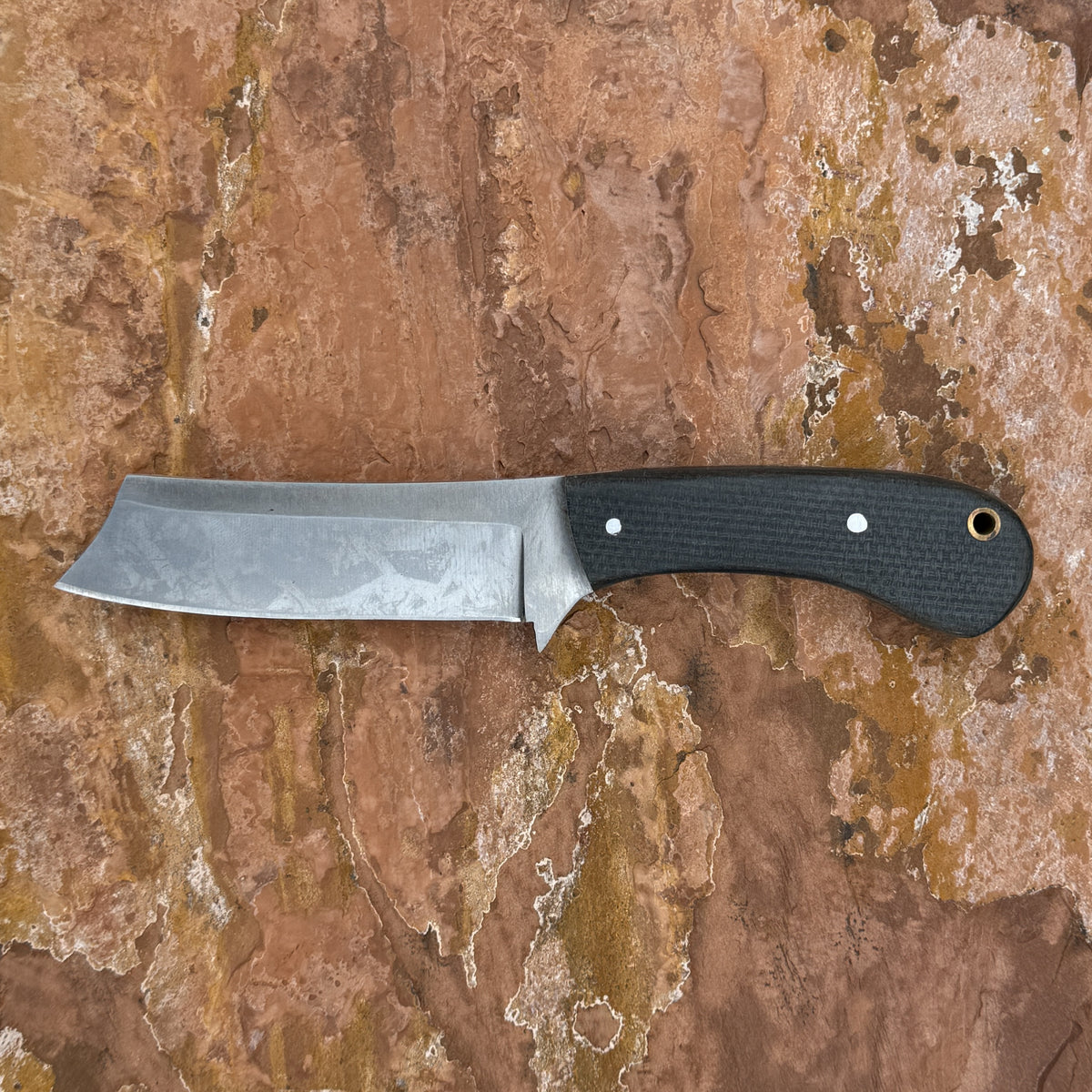 A fixed-blade knife with a satin-finished steel blade and a textured black handle, displayed horizontally on a textured brown and tan stone surface.