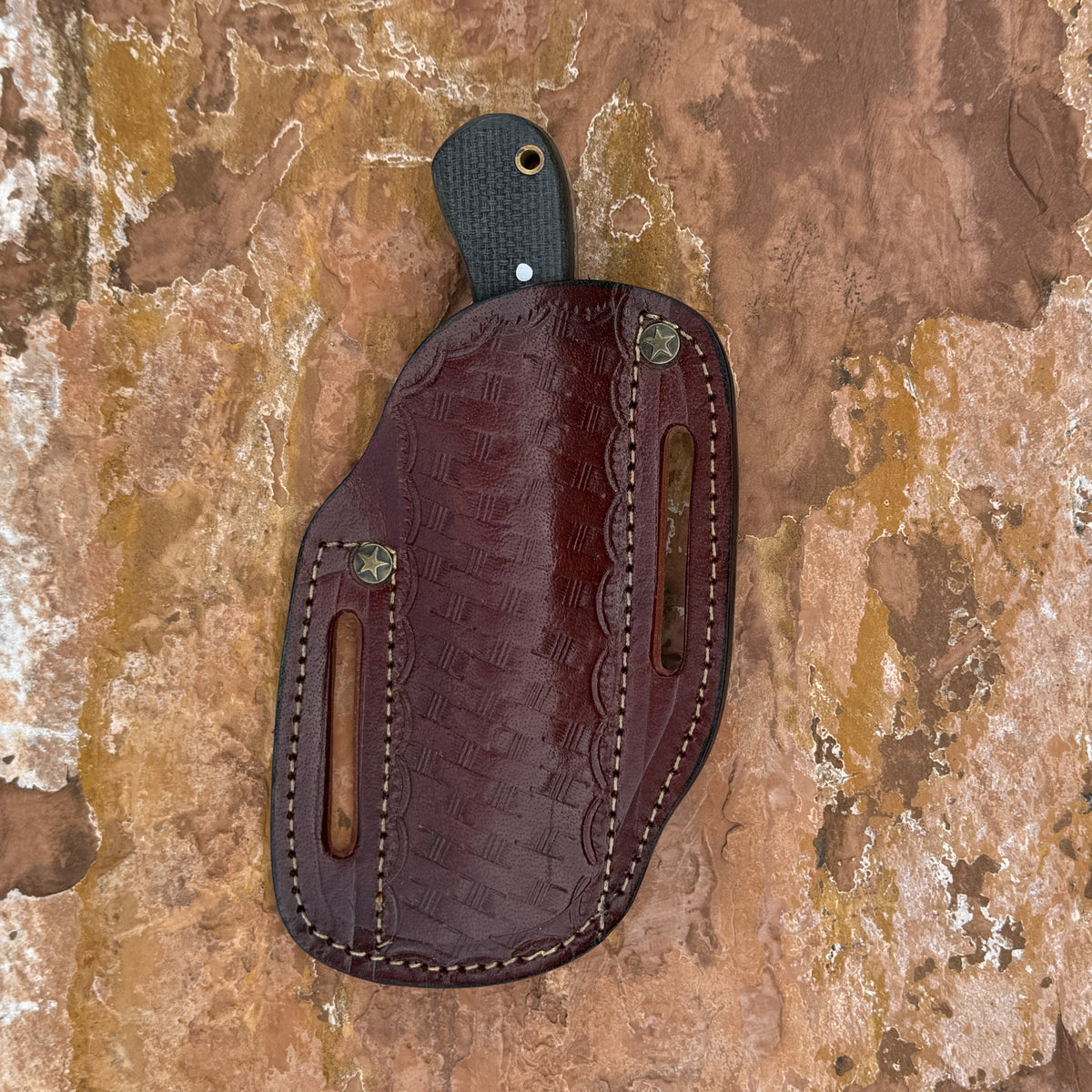 A dark brown embossed leather knife sheath with a textured handle, displayed flat on a textured brown and tan stone surface.