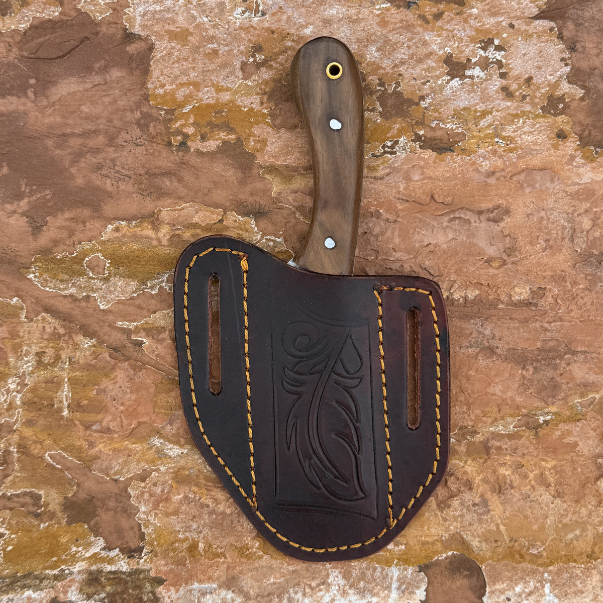 A fixed-blade knife with a dark brown wooden handle and silver pins, resting inside a dark brown leather sheath with tan stitching, set against a textured brown and orange stone background.
