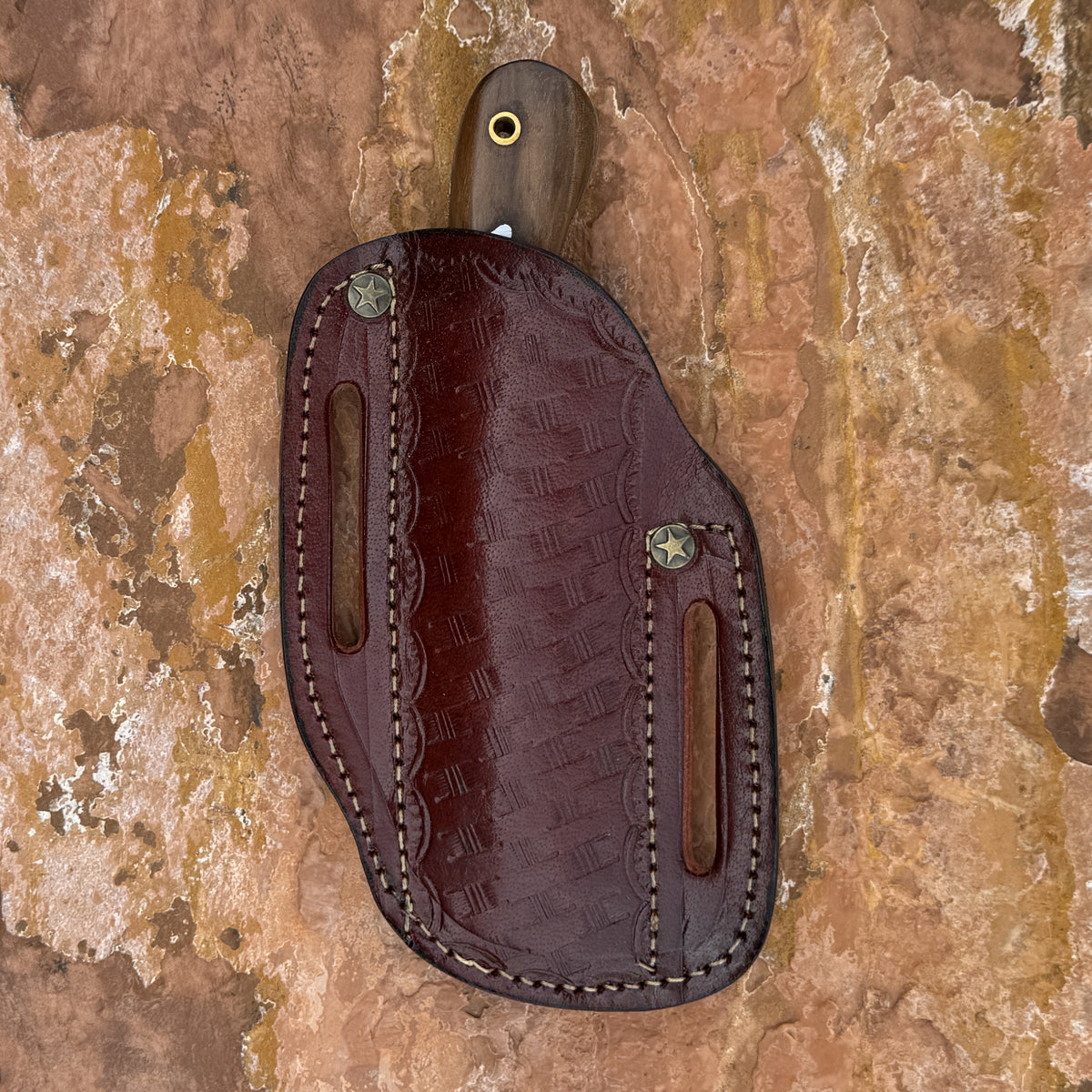 A dark brown leather knife sheath with an embossed crocodile pattern, featuring a wooden handle and a brass eyelet, laid flat on a textured brown and tan stone surface.